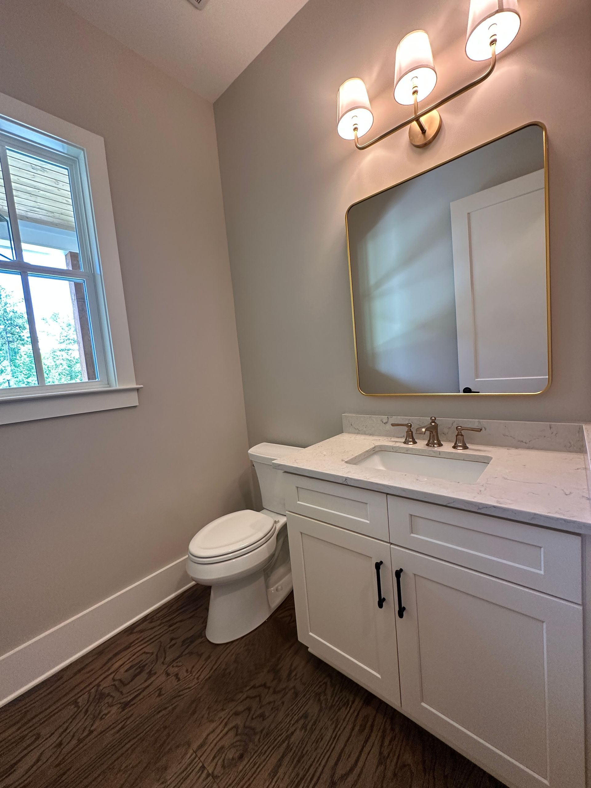 Small bathroom with white vanity, mirror, light fixture, window, and toilet on dark floor.