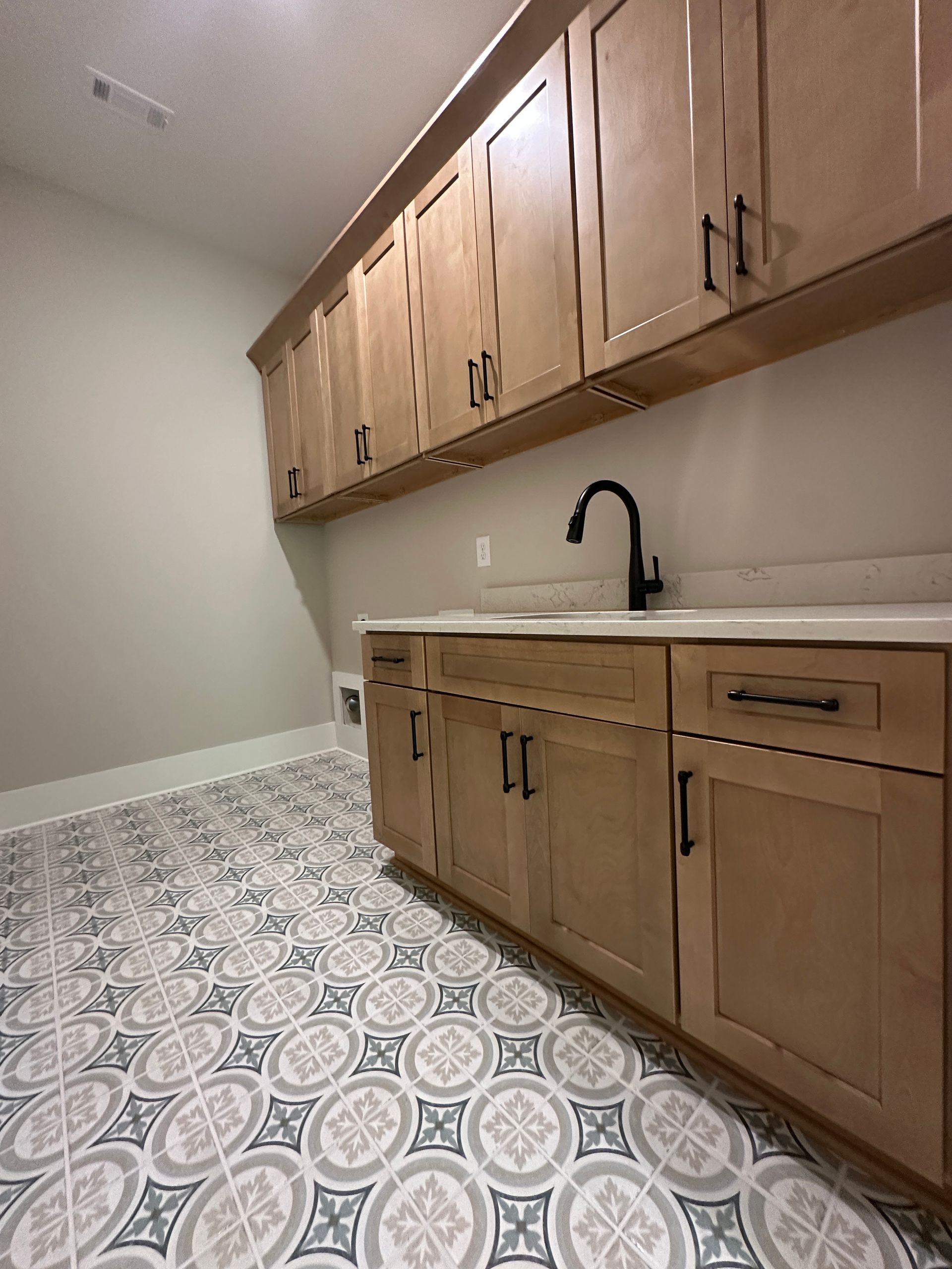 Empty utility room with wood cabinets, white sink, black faucet, and patterned tile floor