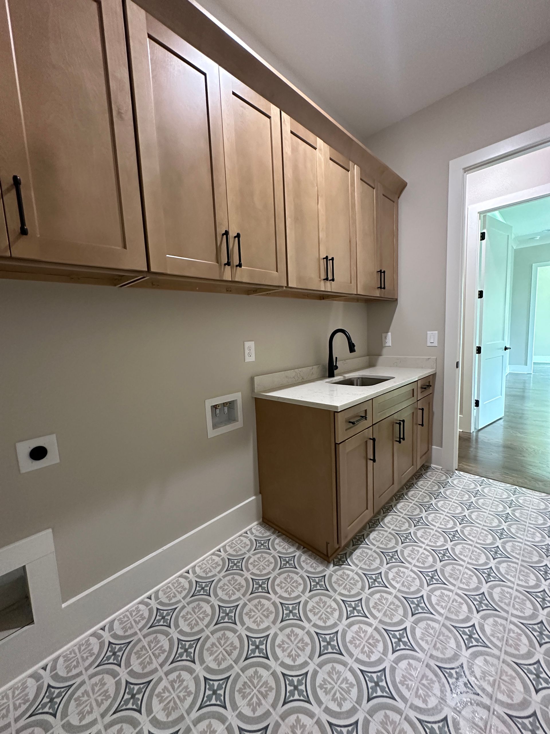 Bright laundry room with wood cabinets, sink, and patterned tile floor leading to a hallway.
