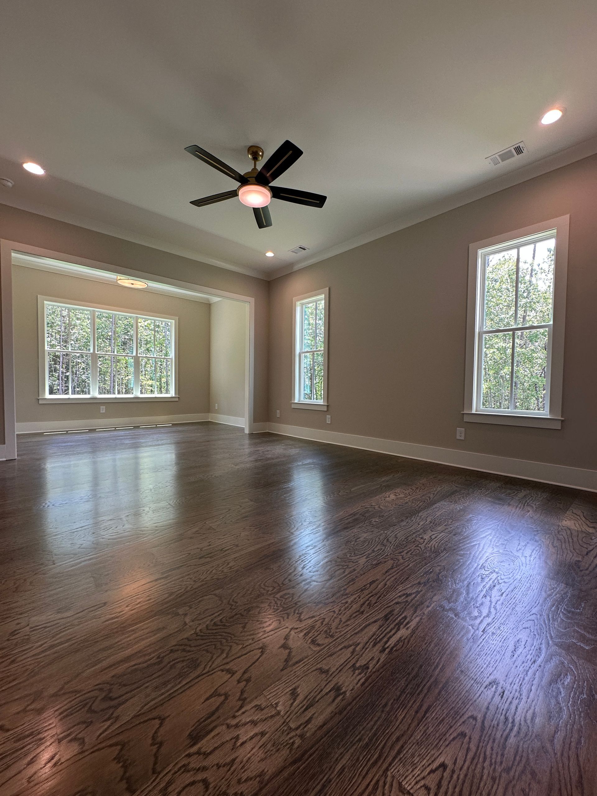 Empty living room with dark hardwood floors, beige walls, and large windows under a ceiling fan.