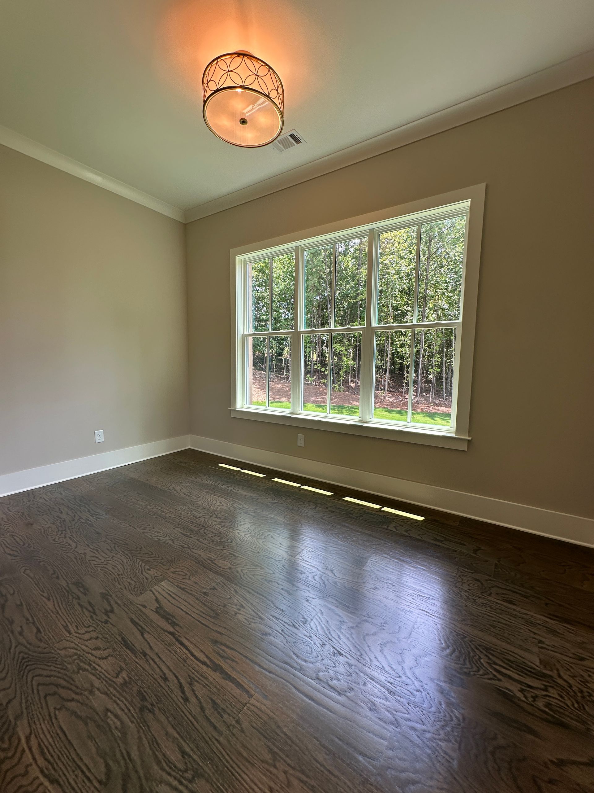Empty room with dark wood floor, beige walls, ceiling light, and large sunlit window.