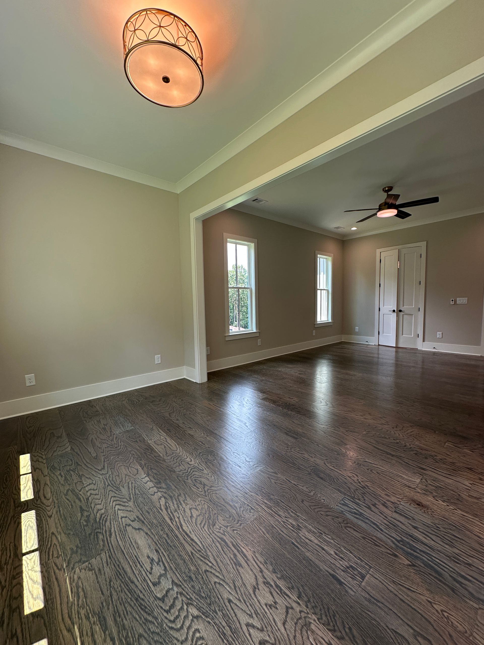 Empty beige living room with dark wood floors, ceiling fan, and large windows with white trim
