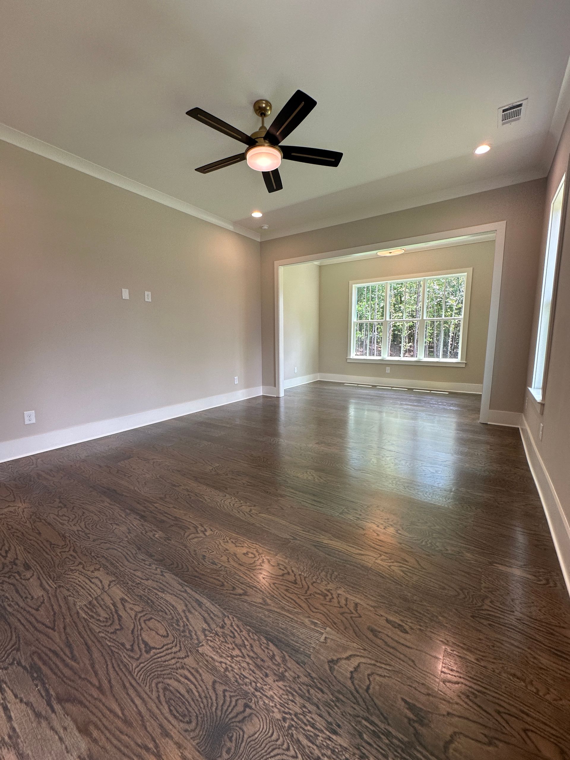 Empty beige living room with dark hardwood floors, ceiling fan, and large windows at the far end