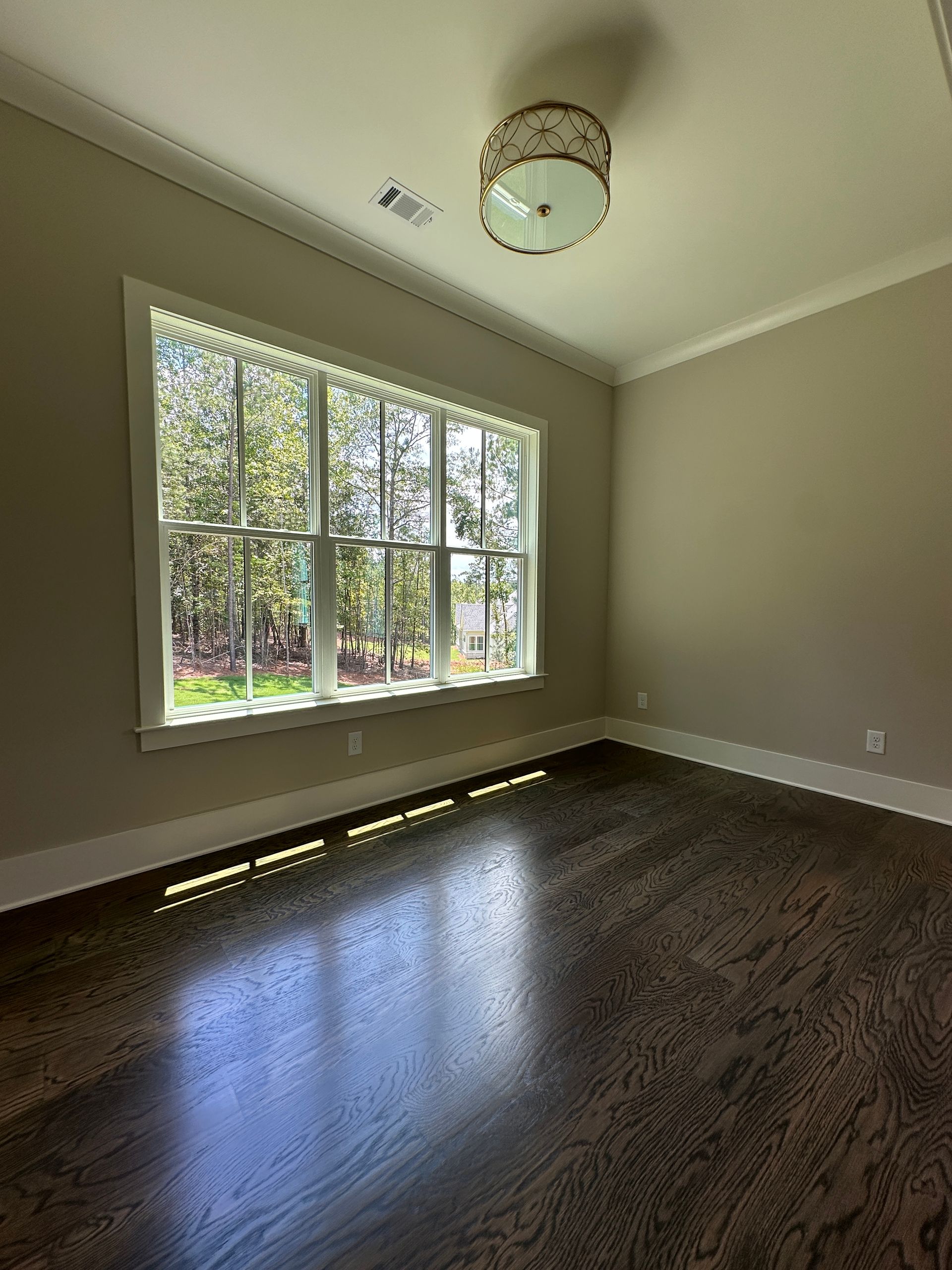 Empty sunlit room with large window, beige walls, dark hardwood floor, and ceiling light fixture
