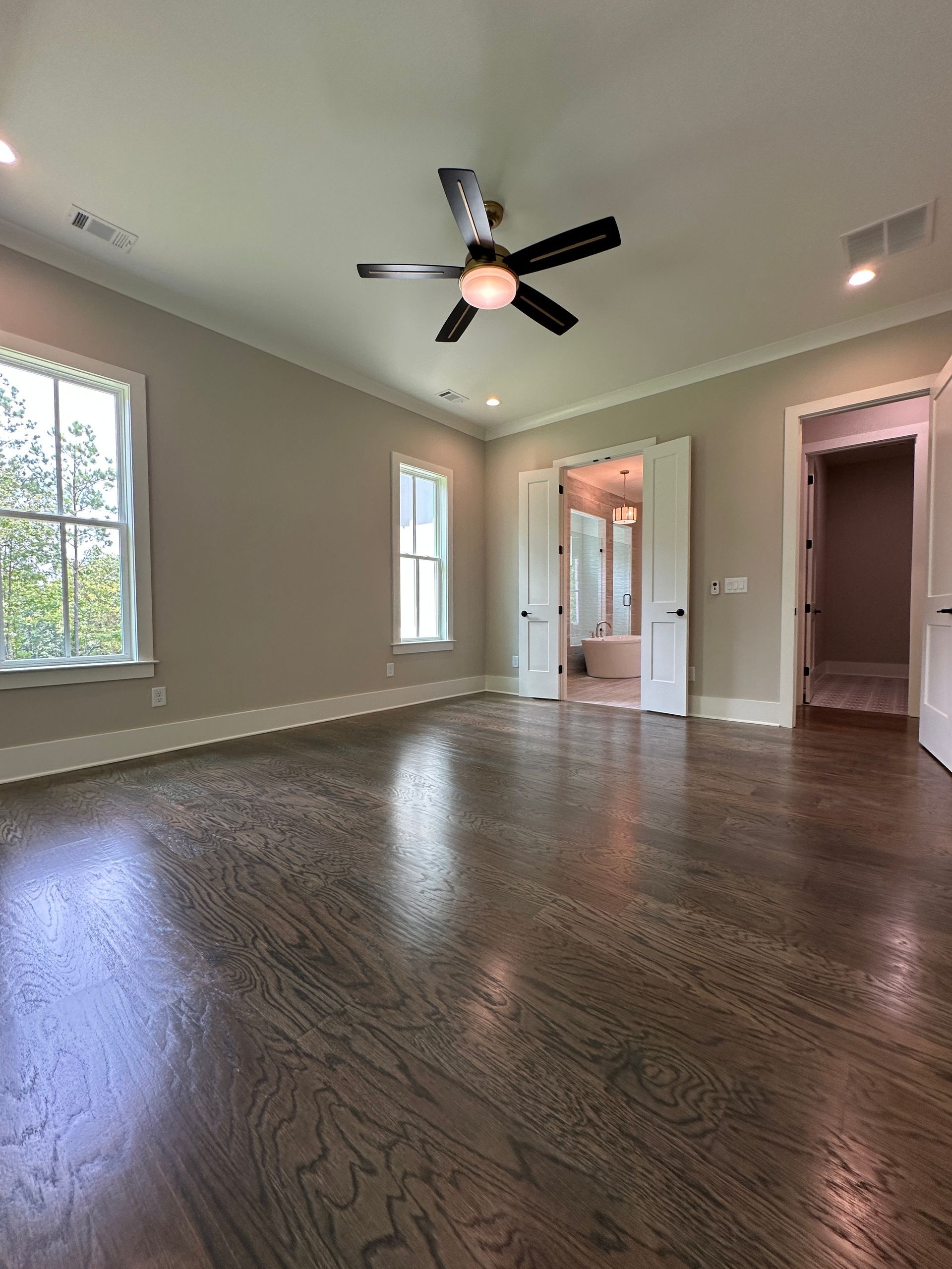 Empty carpeted living room with ceiling fan, large windows, and adjacent hall and bathroom doorways