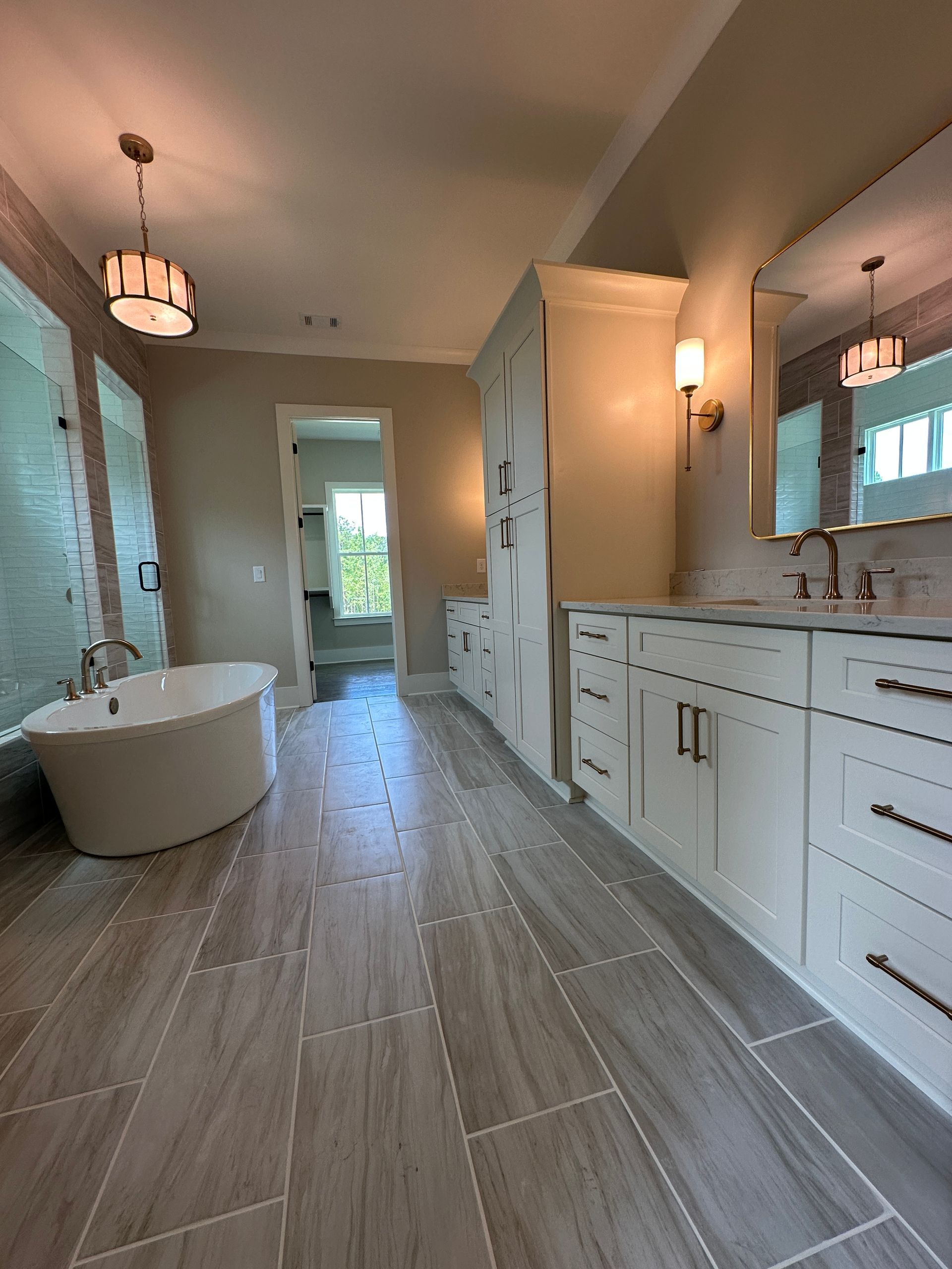 Modern bathroom with freestanding tub, double vanity, and long gray tiled floor leading to a bright doorway