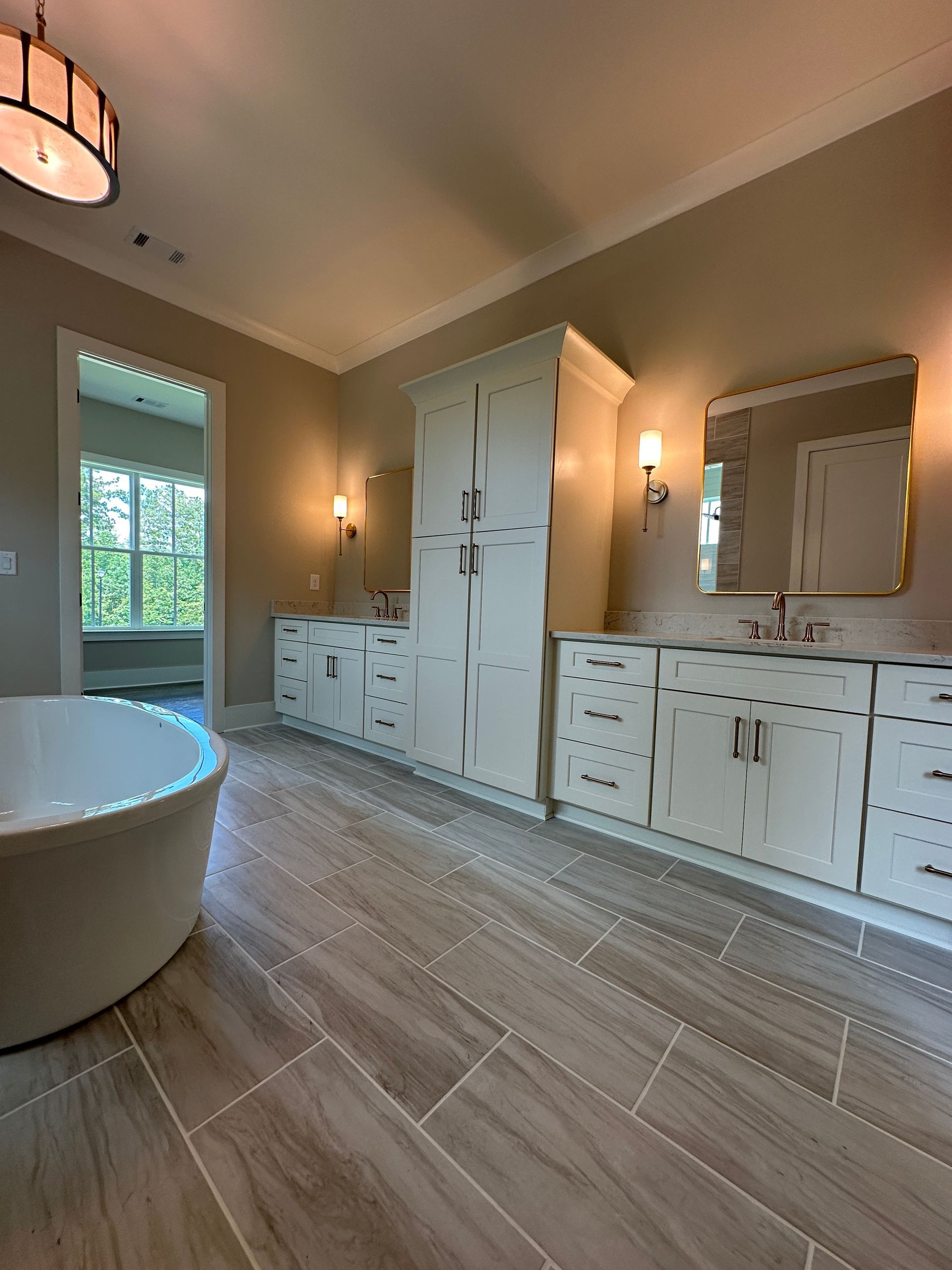 Bright bathroom with freestanding tub, white cabinets, mirror, sconces, and wood-look tile floor.