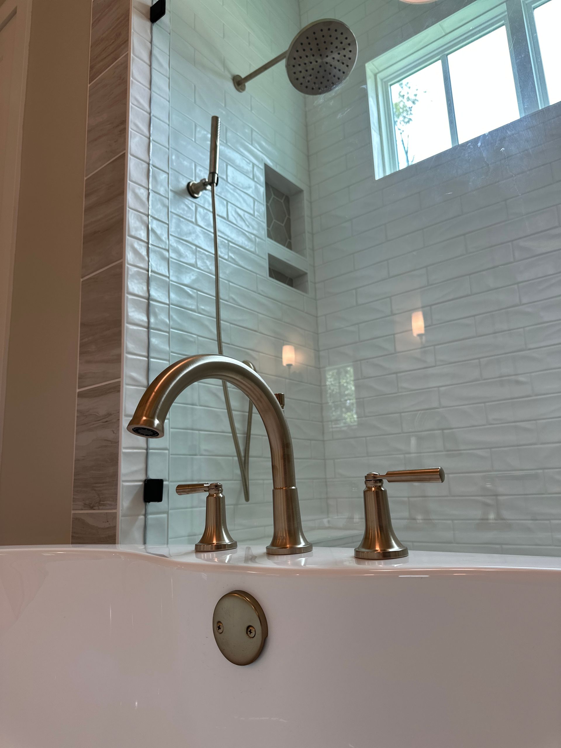 Bright bathroom with white tiled shower, window, and a bathtub faucet in the foreground