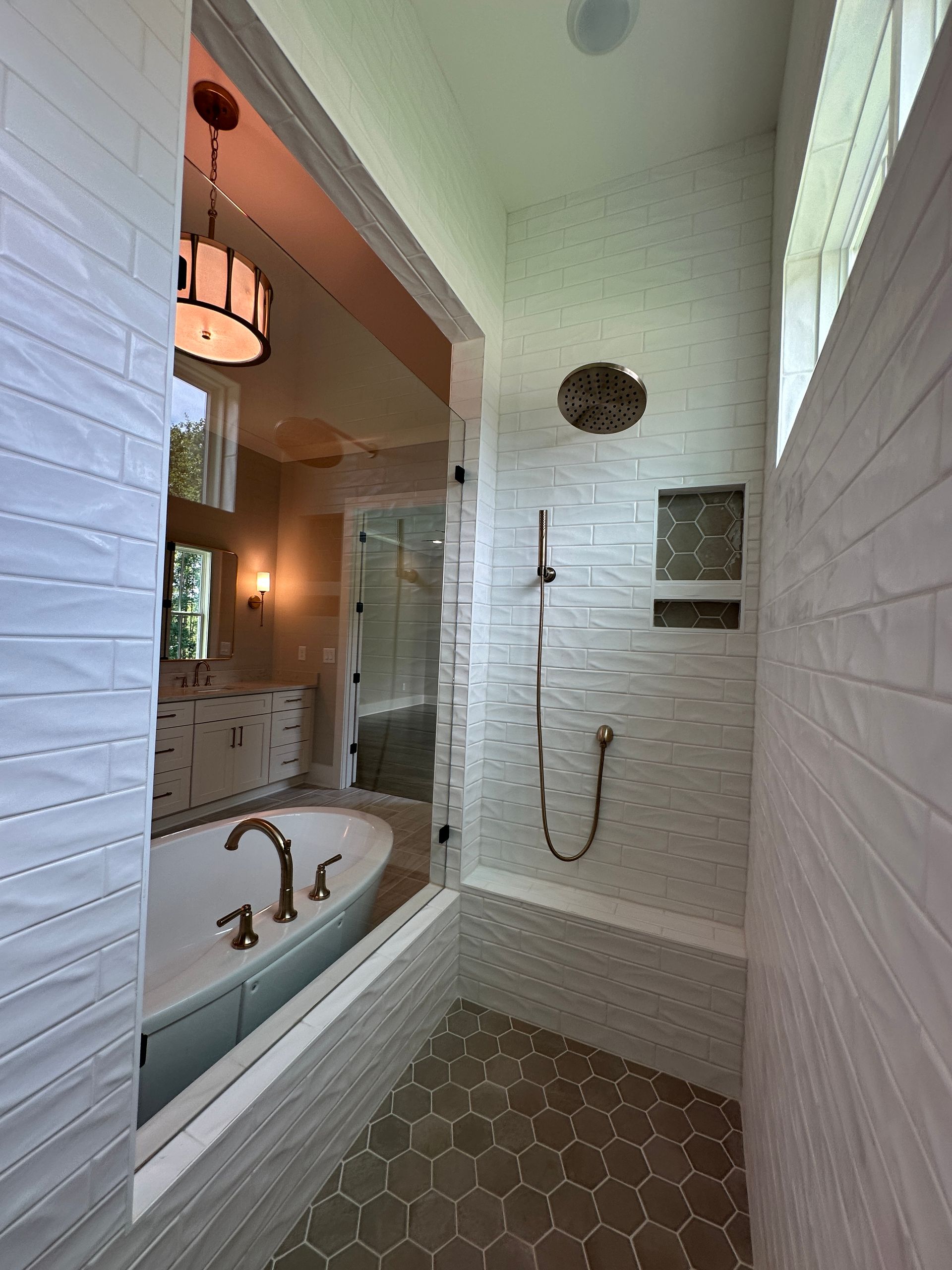 Bright bathroom with white subway tile shower, hexagon floor, tub, and wall mirror reflected on the left.
