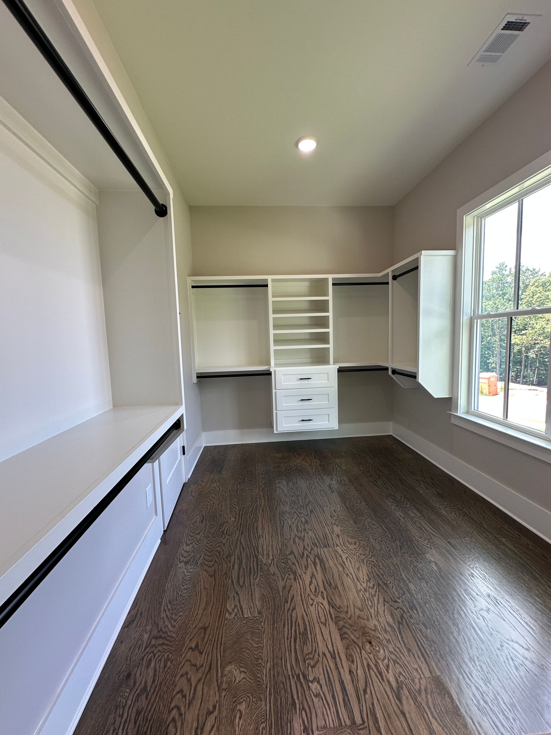 Empty walk-in closet with dark wood floor, white built-in shelving, and a window letting in daylight