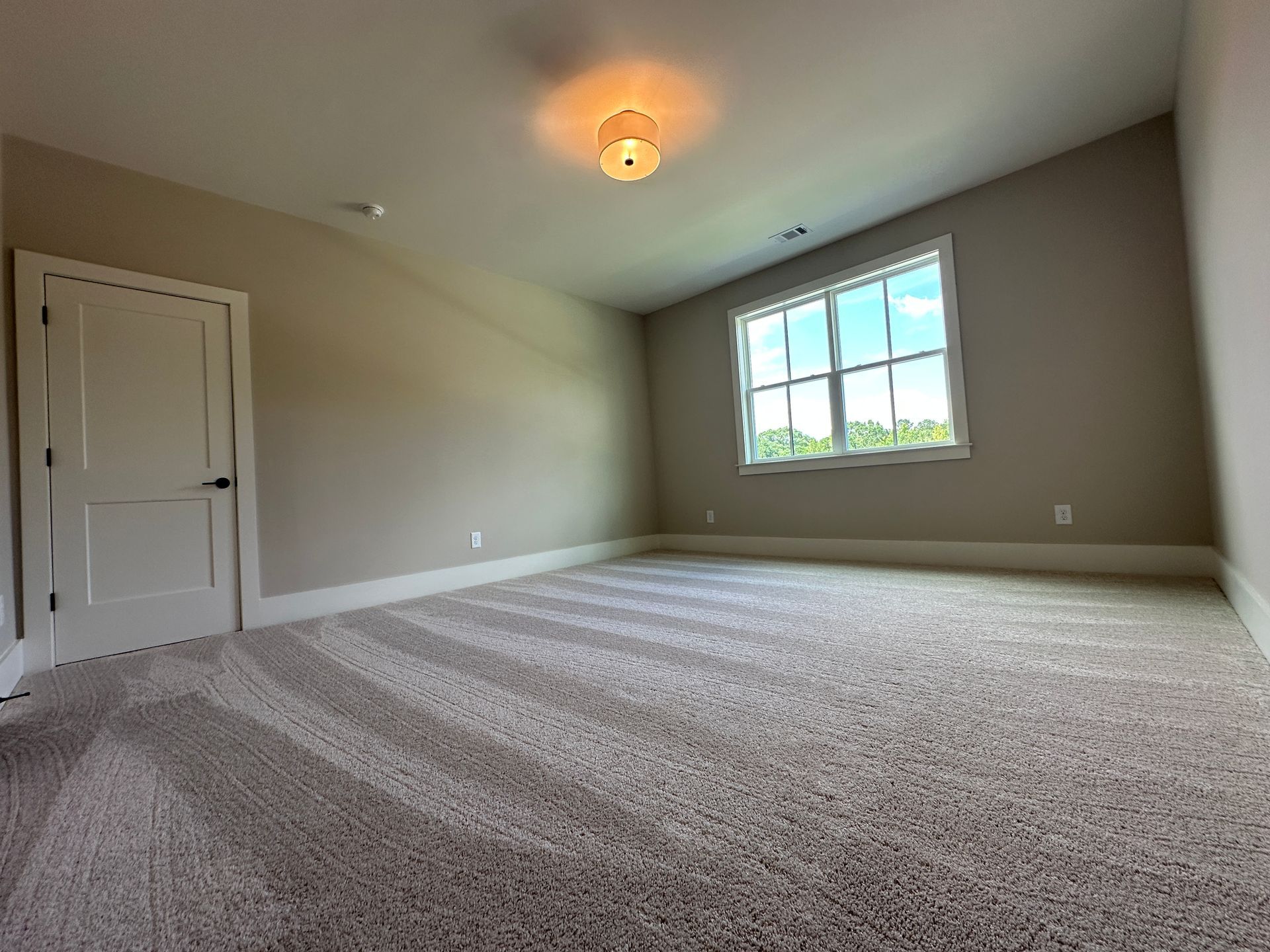 Empty beige bedroom with carpet, white closet doors, and a large window letting in daylight
