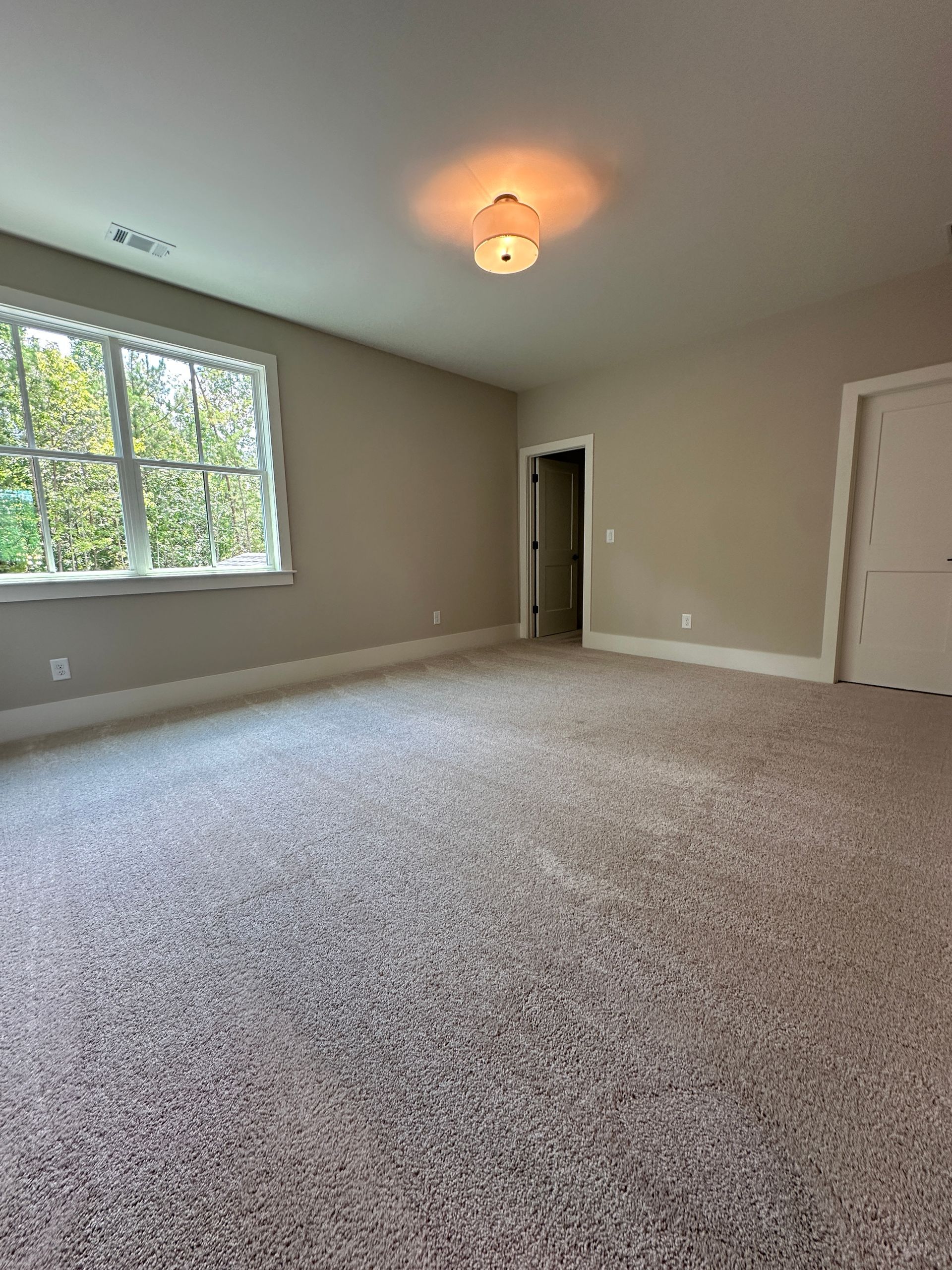 Empty beige bedroom with carpet, window, ceiling light, and open doorway