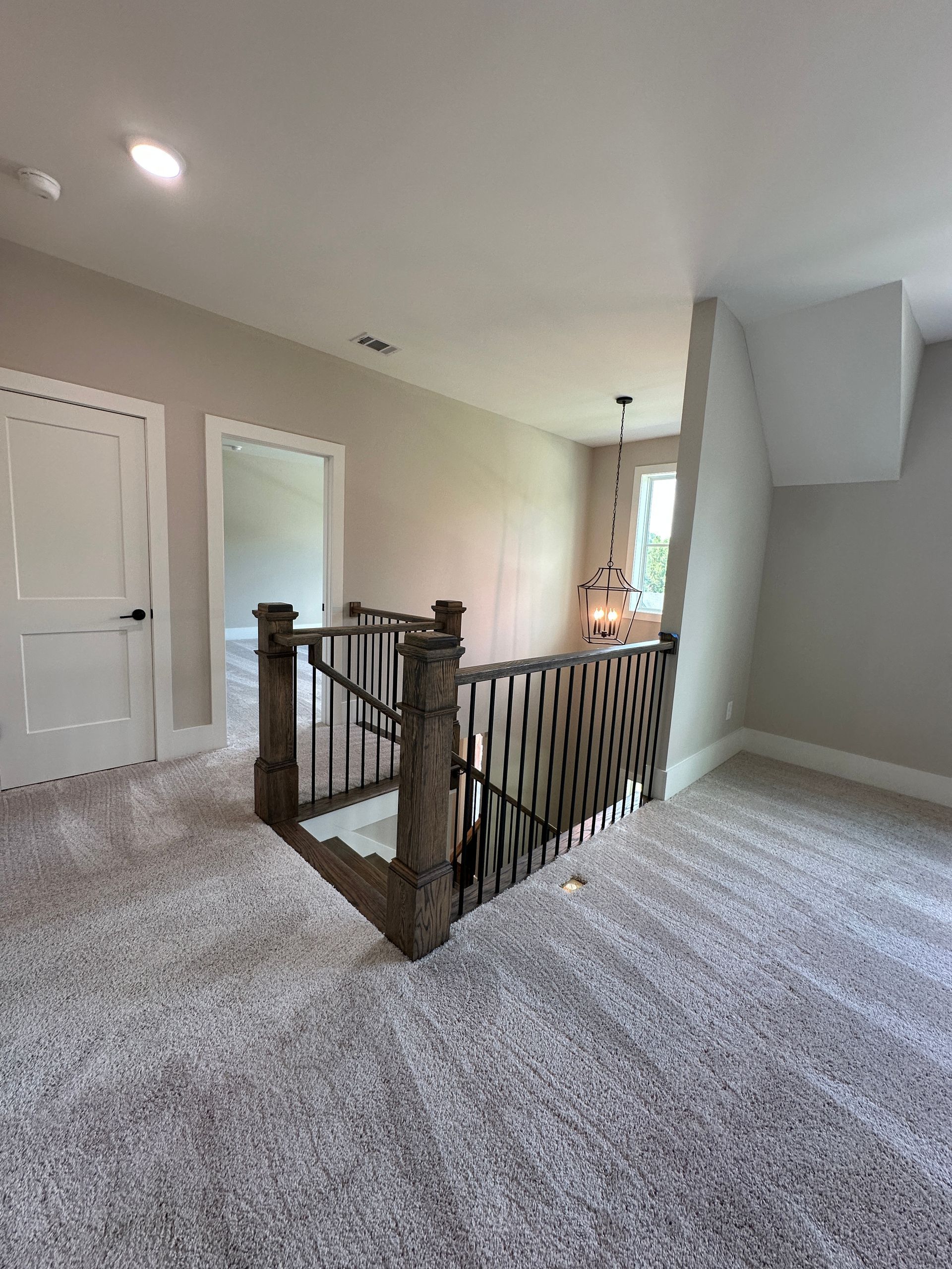 Carpeted upstairs hallway with wooden railing, white walls, and a view to a bright window.
