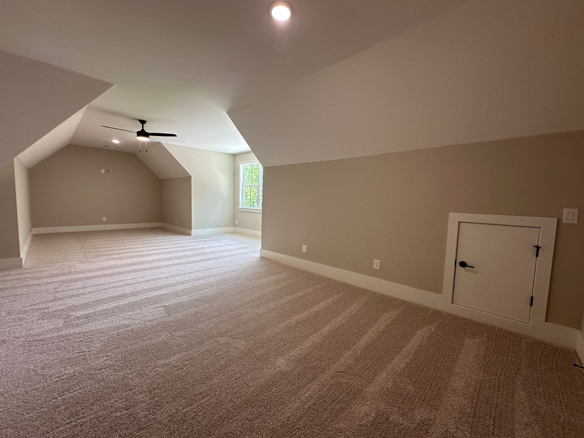 Empty beige carpeted attic room with sloped ceiling, ceiling fan, and small window.