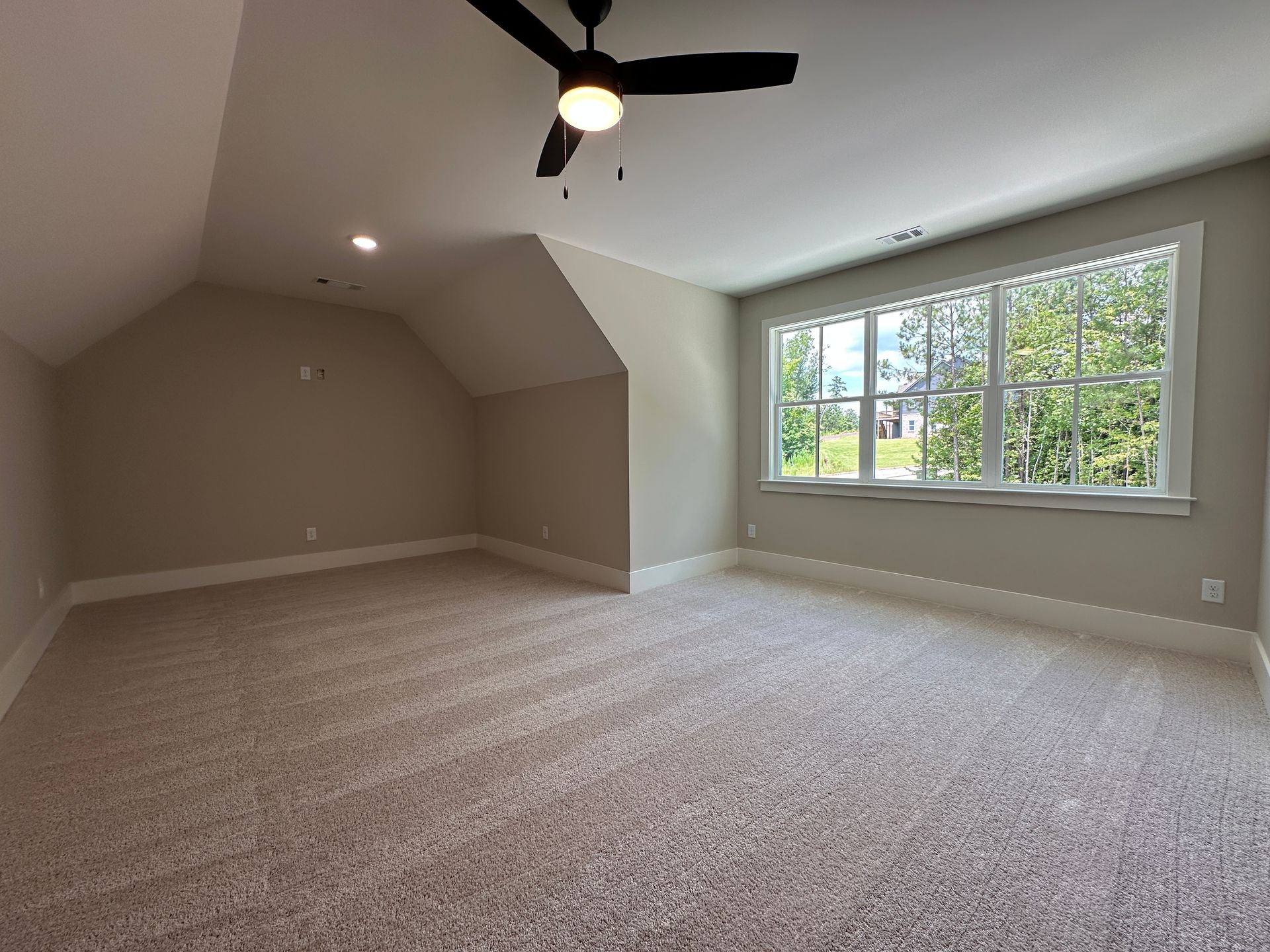 Empty beige carpeted bedroom with sloped ceiling, black ceiling fan, and a large window.