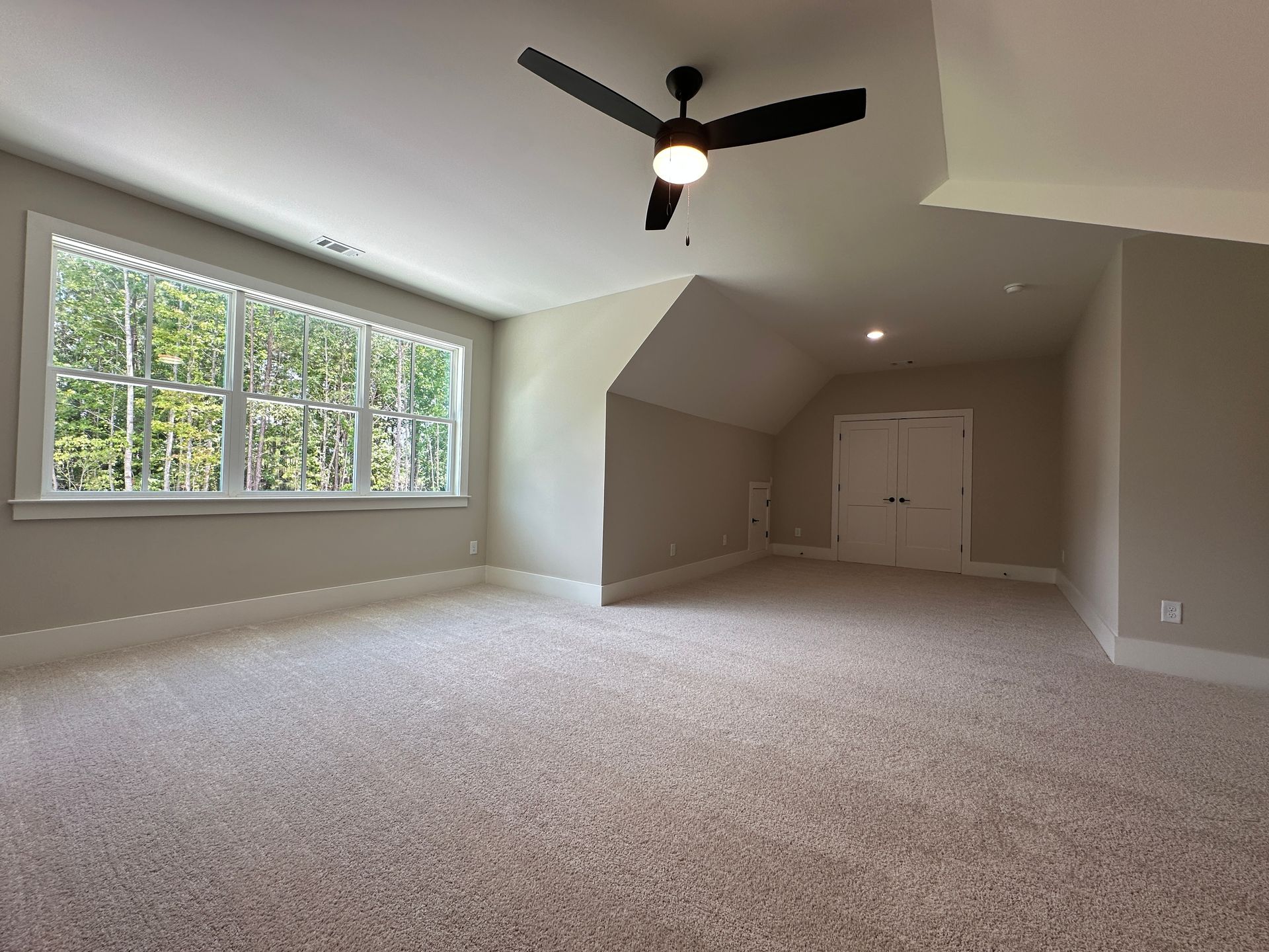 Empty carpeted living room with large window, vaulted ceiling, ceiling fan, and double doors