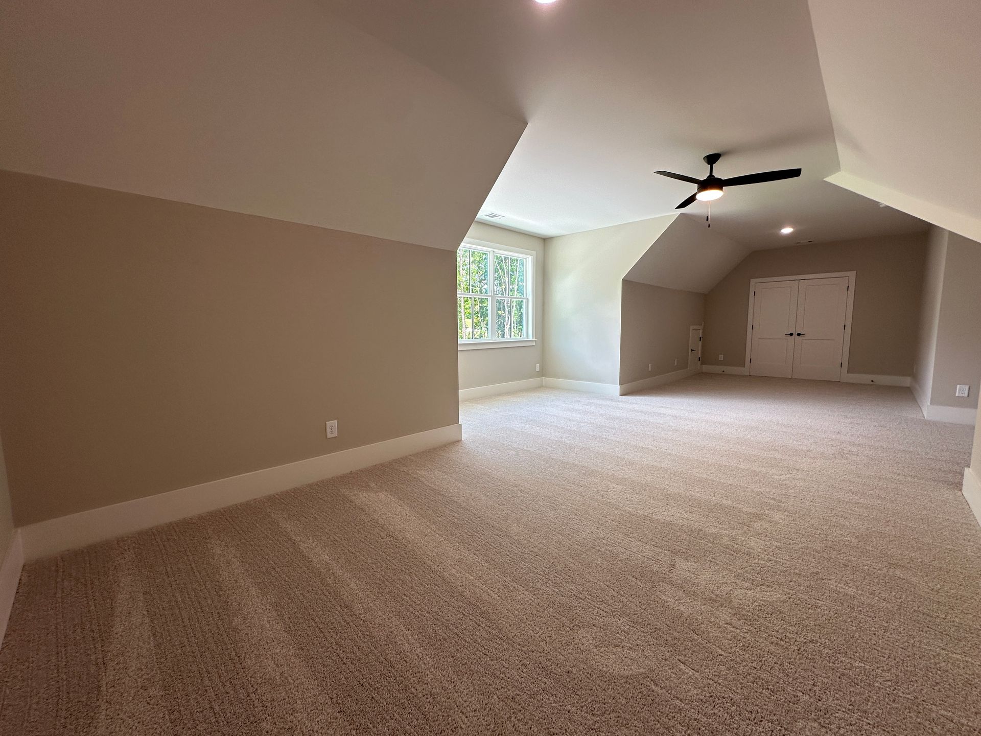 Empty beige carpeted room with sloped ceiling, ceiling fan, and a window at the far end.