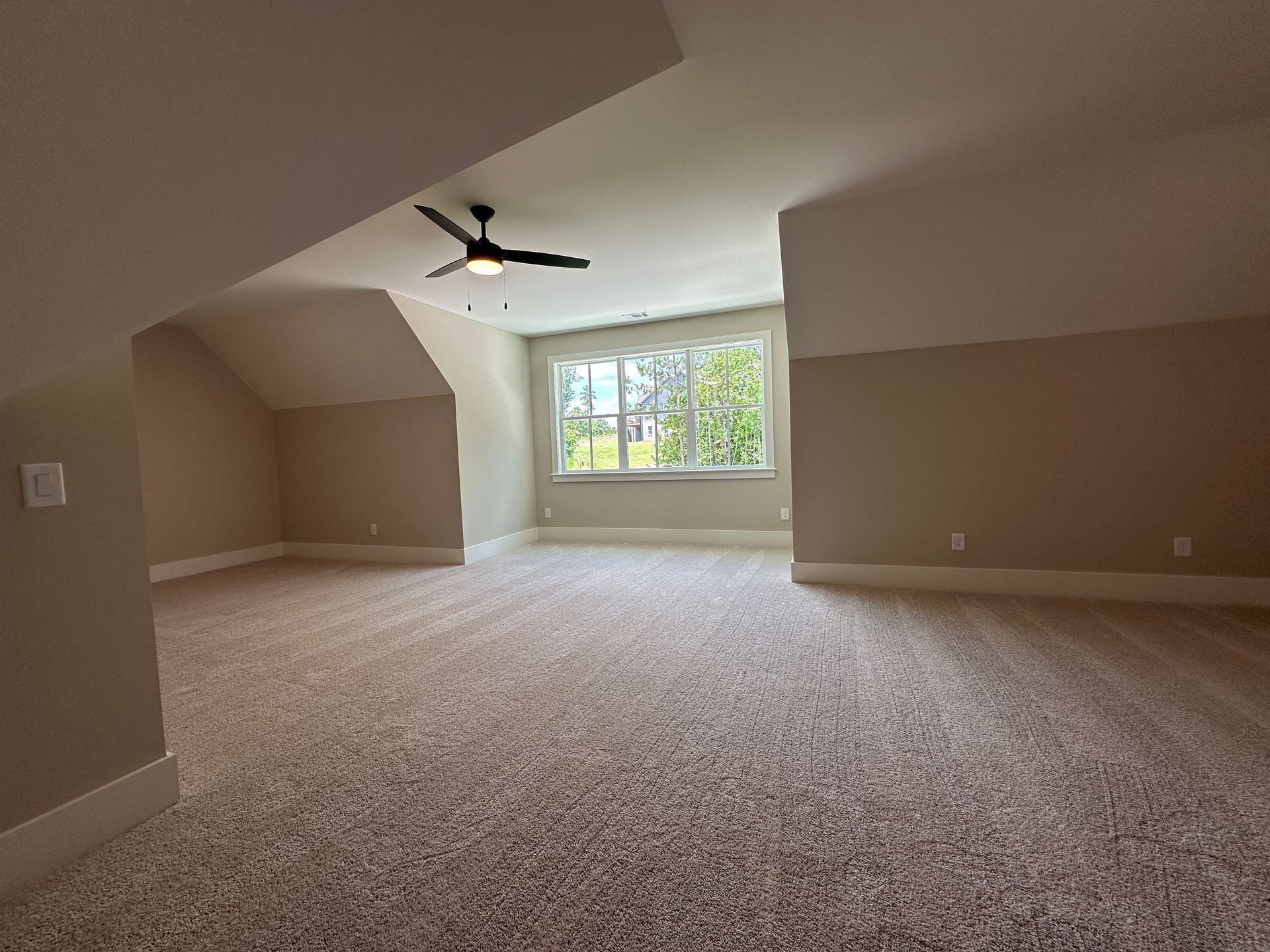 Empty beige bedroom with carpeted floor, sloped ceiling, ceiling fan, and large window letting in daylight