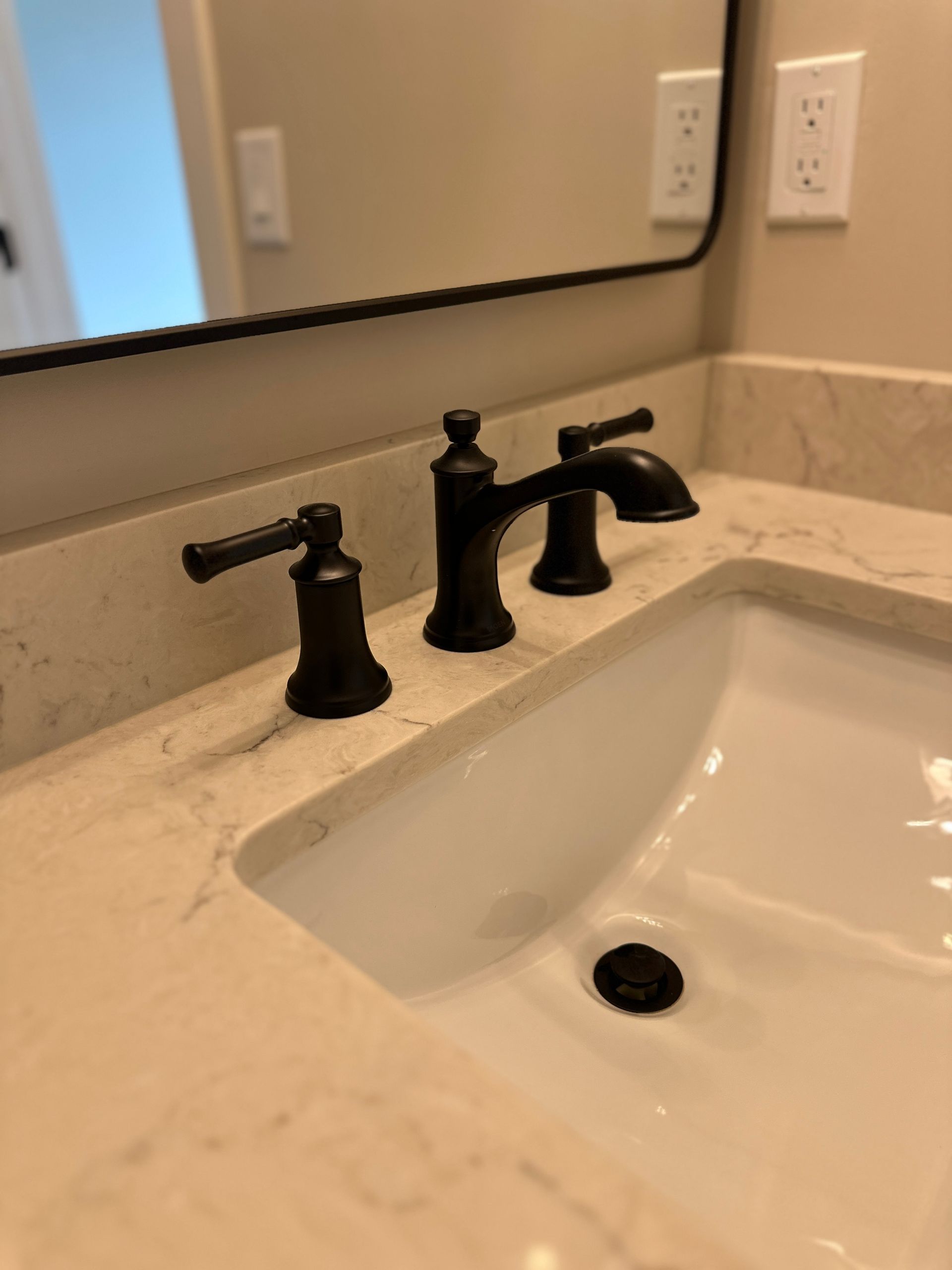 Bathroom sink with black faucet and handles on a light countertop, seen in a mirror.