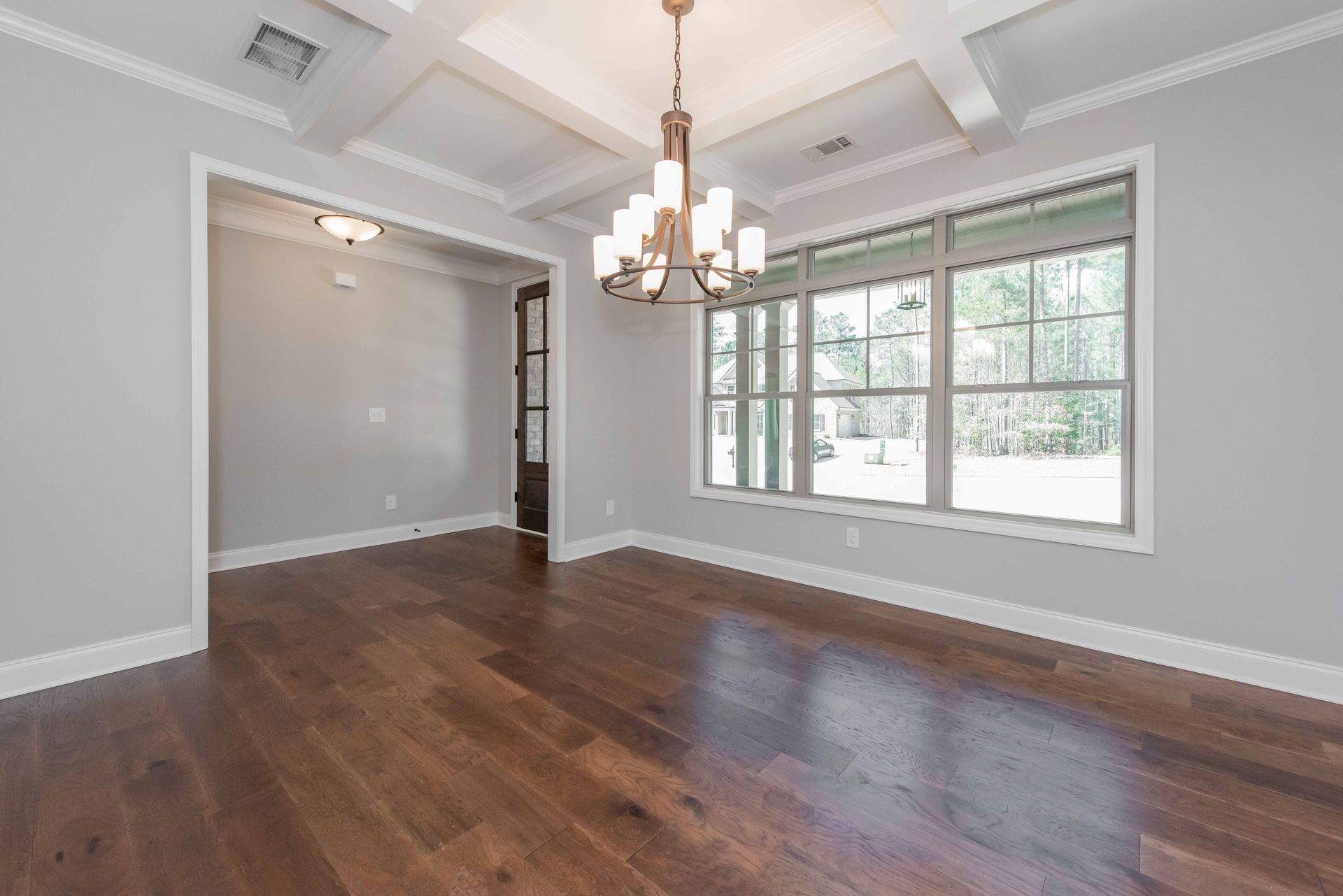 Empty living room with dark hardwood floors, white walls, chandelier, and large front windows