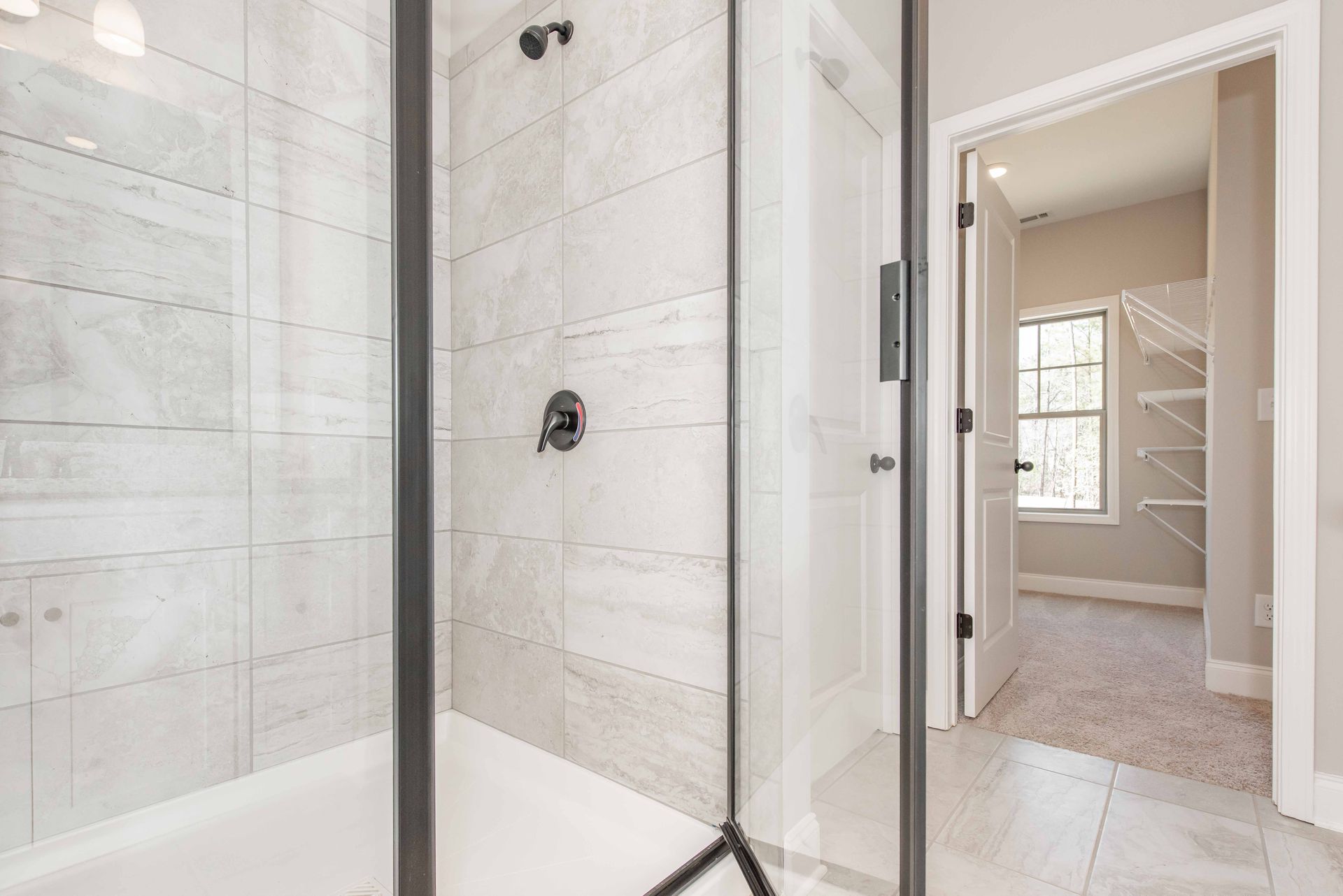 Modern tiled bathroom with glass-enclosed shower and open door to a carpeted closet area