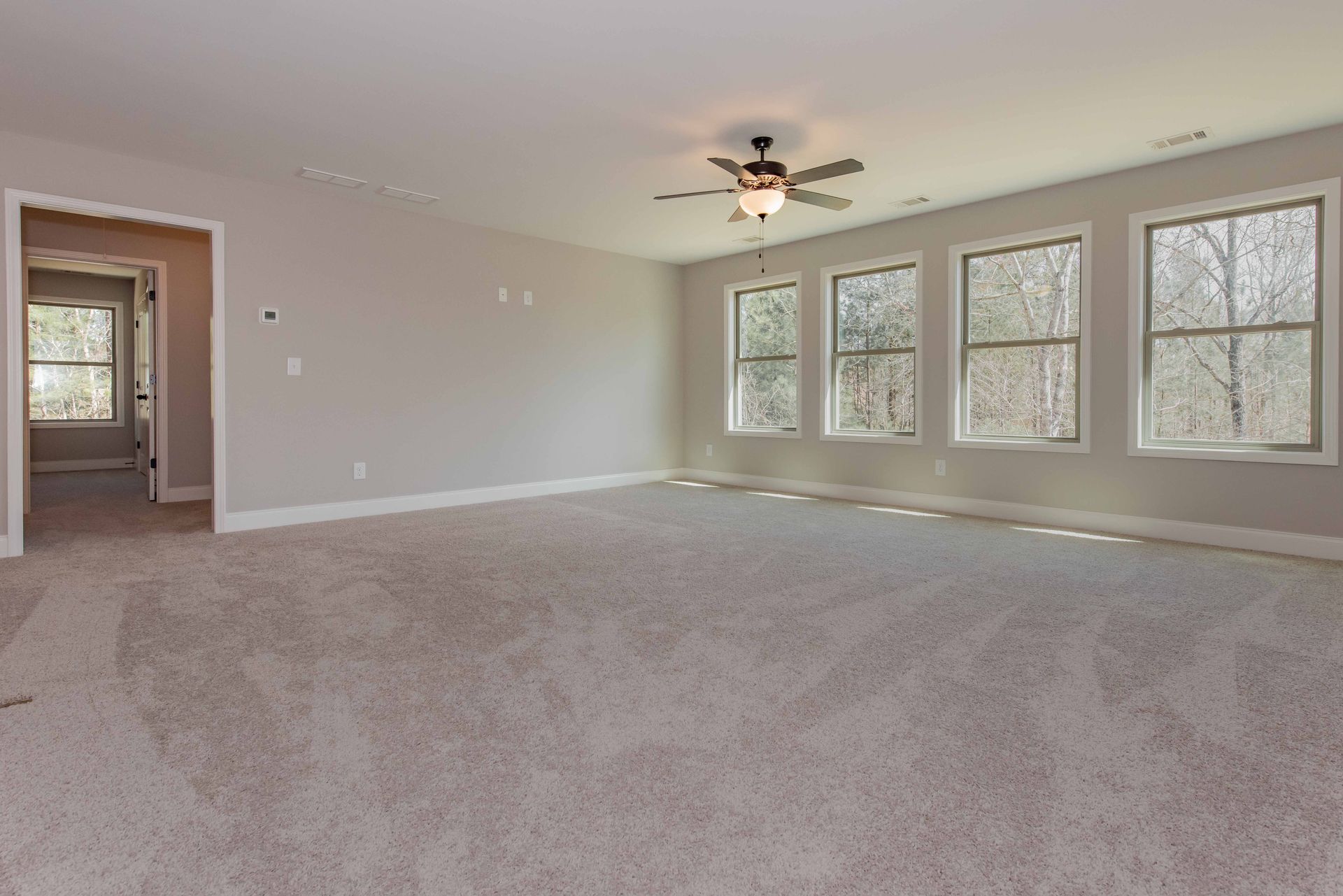 Empty carpeted living room with gray walls, ceiling fan, and large windows.