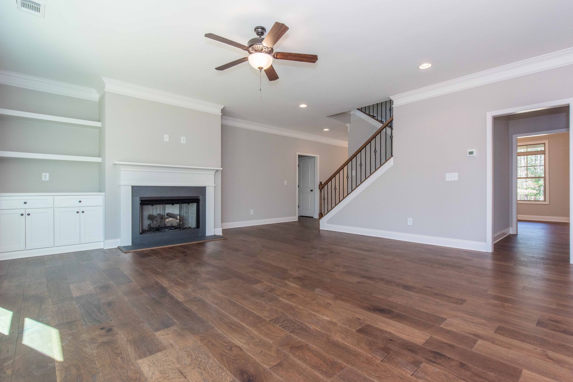Empty living room with dark wood floors, fireplace, ceiling fan, staircase, and open doorway to a kitchen