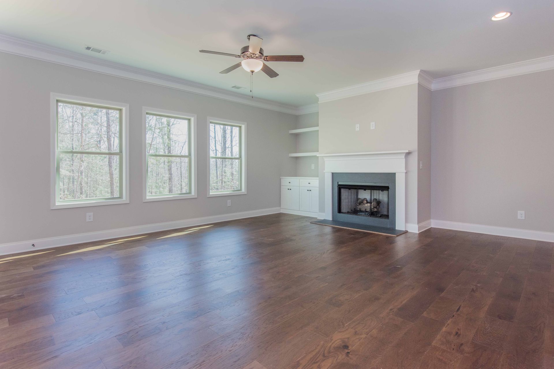 Empty living room with hardwood floors, large windows, a ceiling fan, and a fireplace.