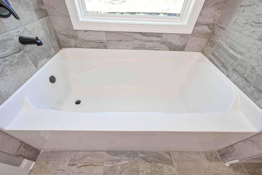 White bathtub with gray tiled walls, below a window.
