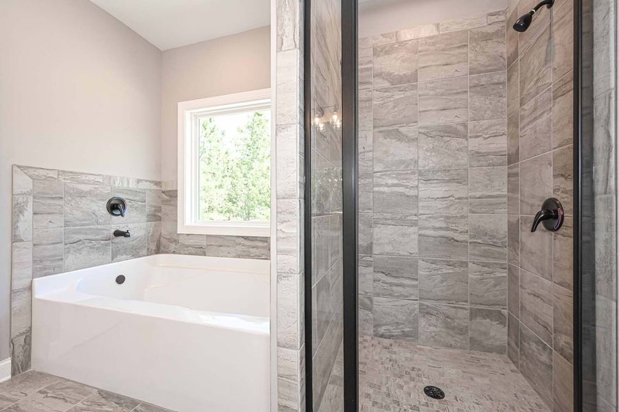 Bathroom with a white bathtub, a glass-door shower, grey tiled walls, and a window.