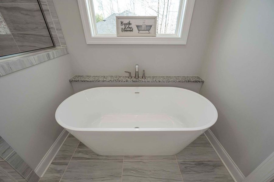 White oval soaking tub in a gray-tiled bathroom with window and countertop.