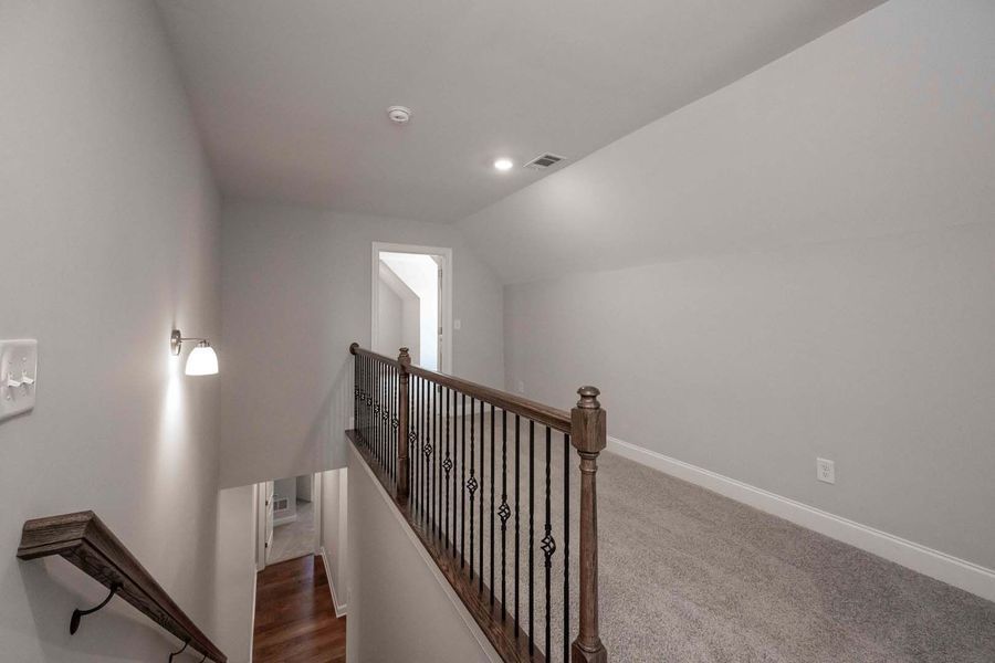 A gray hallway with a stairway, carpeted floor, and a dark brown railing.