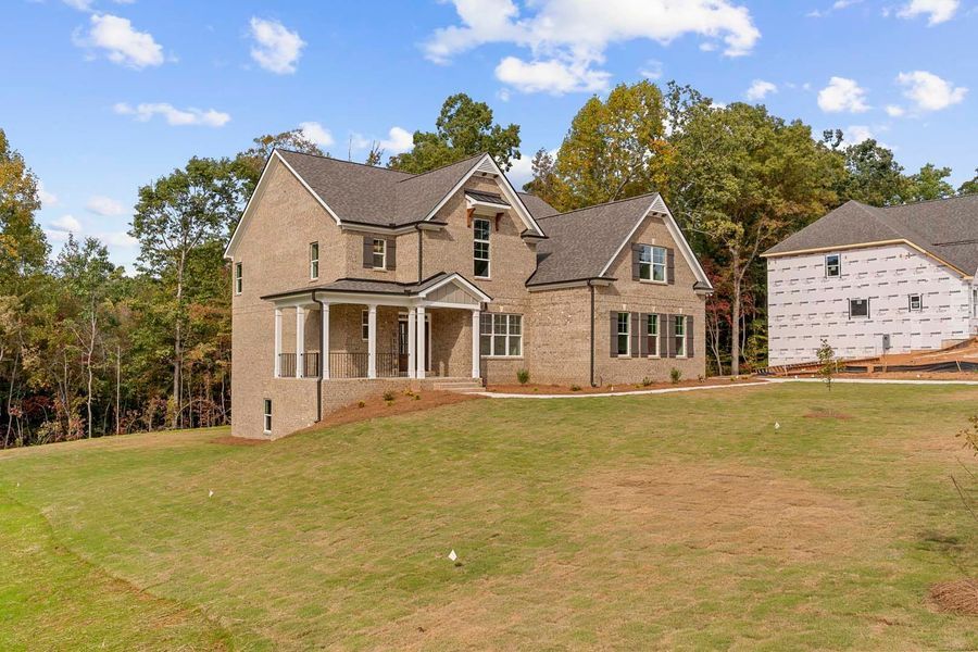 Two-story brick house with a porch on a grassy hill, surrounded by trees, and another house under construction.
