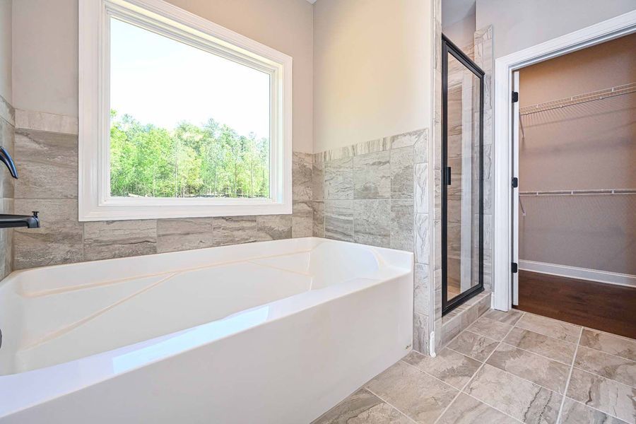 Bathroom with a white tub under a window. Stone tile lines the walls and floor. A glass shower is nearby.