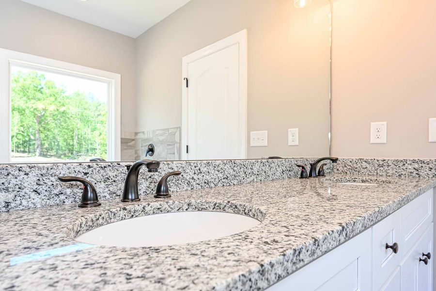 Bathroom with granite countertop, double sinks, bronze fixtures, white cabinets, large mirror, and window.