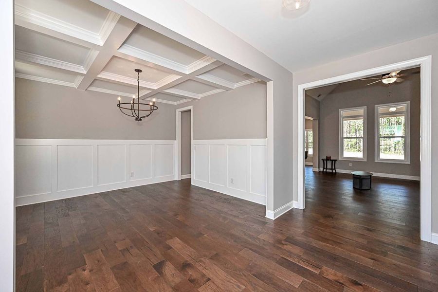 Empty dining room with wainscoting, coffered ceiling, and dark wood floor. Open doorway leads to living area.
