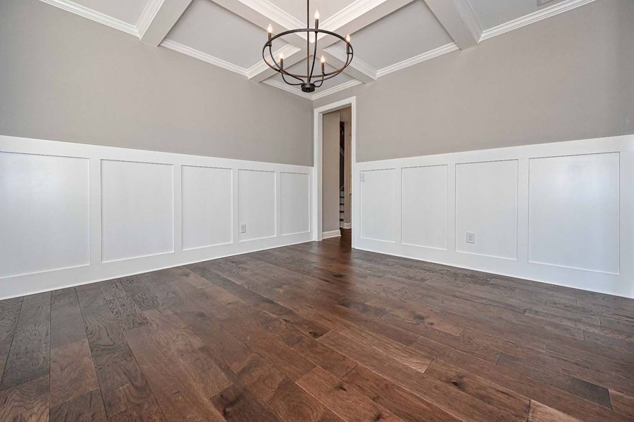 Empty room with wood floors, white paneling, and gray walls, chandelier hangs from the coffered ceiling.