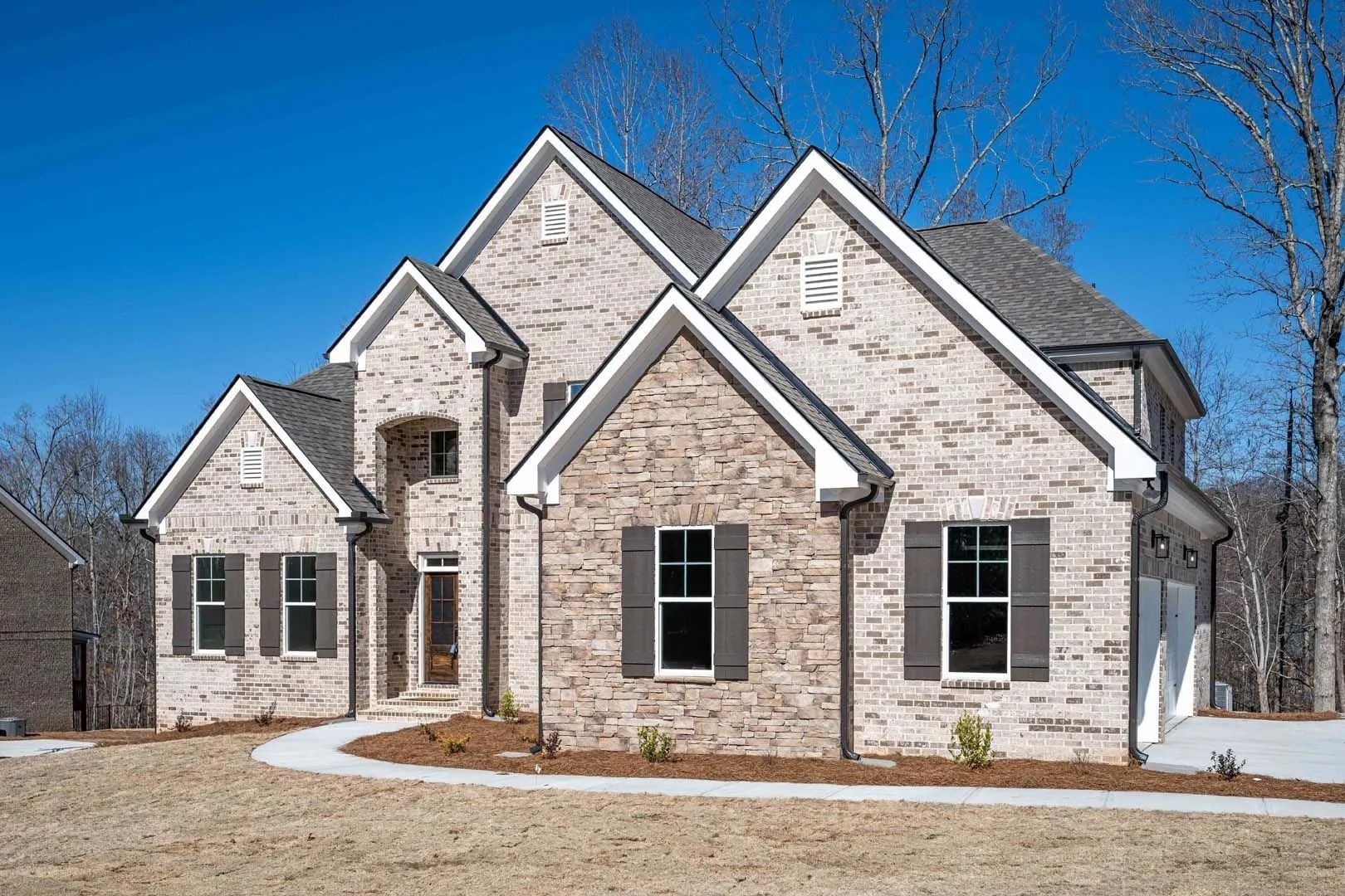 Two-story brick house with gray roof, dark shutters, and blue sky in a sunny setting.