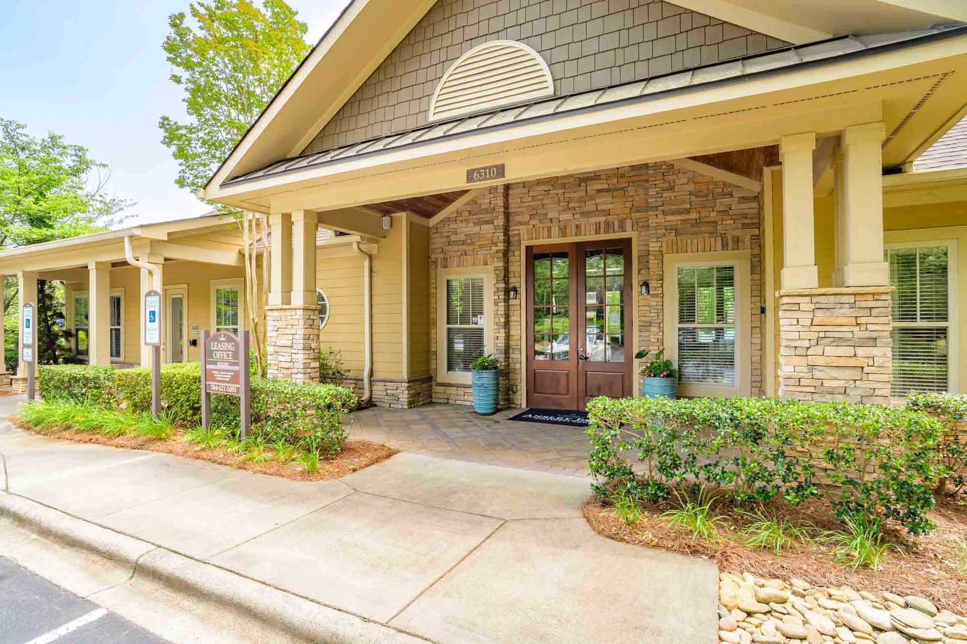 Exterior of a tan and brick building with columns, a brown door, and landscaping at Ansley Falls Apartment Homes in Charlotte, NC.
