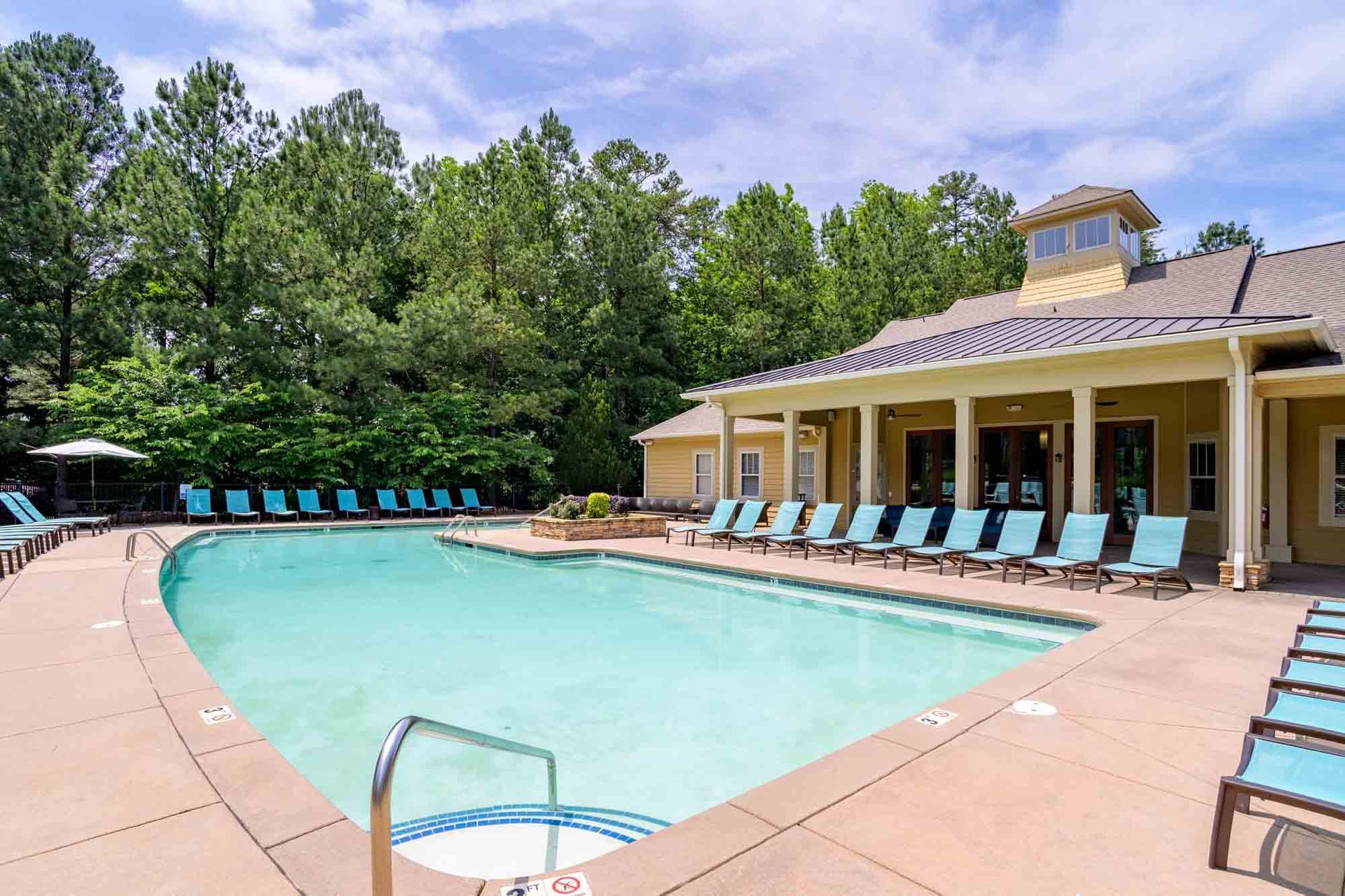 Swimming pool with blue lounge chairs, next to a building and trees on a sunny day at Ansley Falls Apartment Homes in Charlotte, NC.