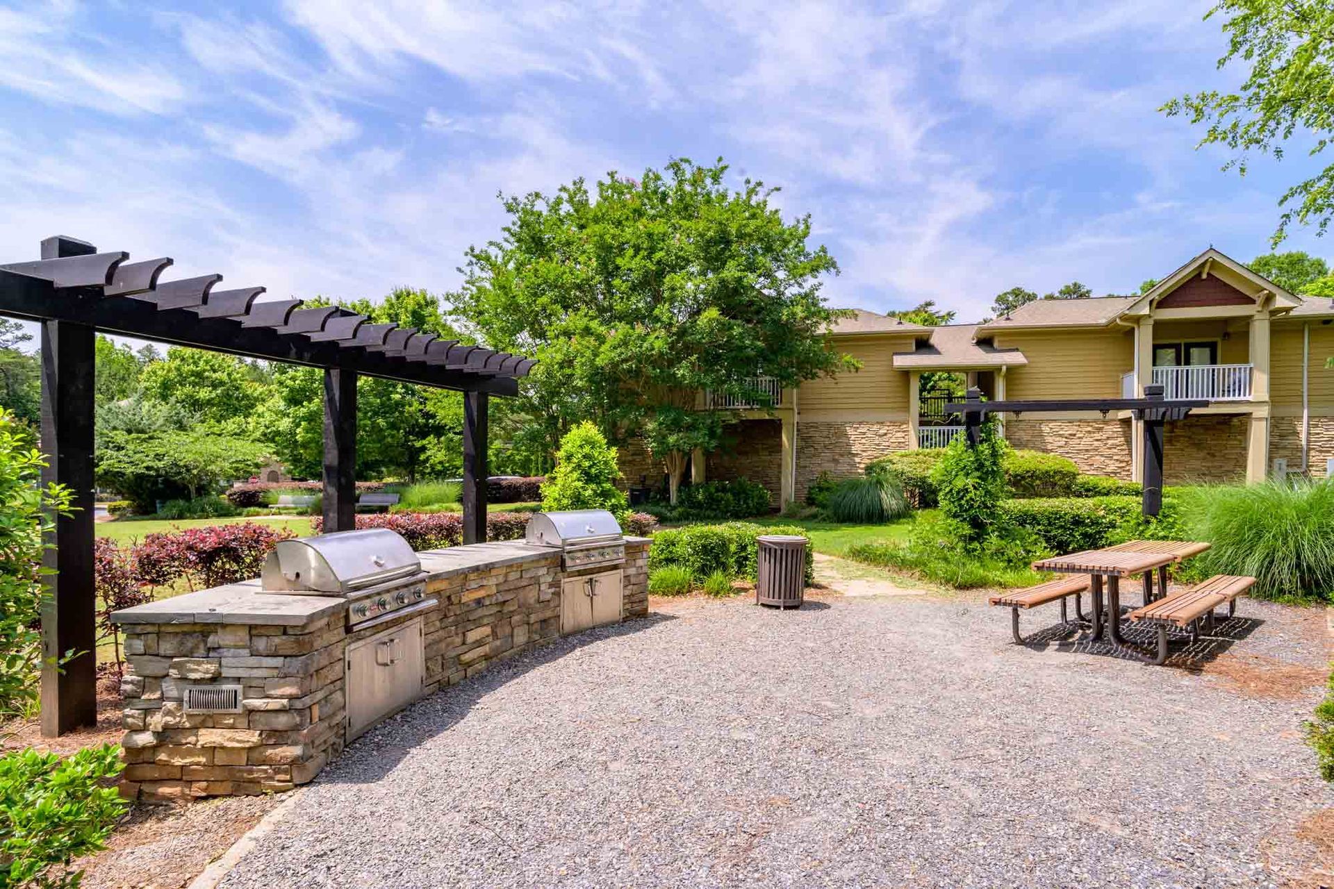 Outdoor grilling area with stone grill, pergola, picnic table, and apartment building in background at Ansley Falls Apartment Homes in Charlotte, NC.