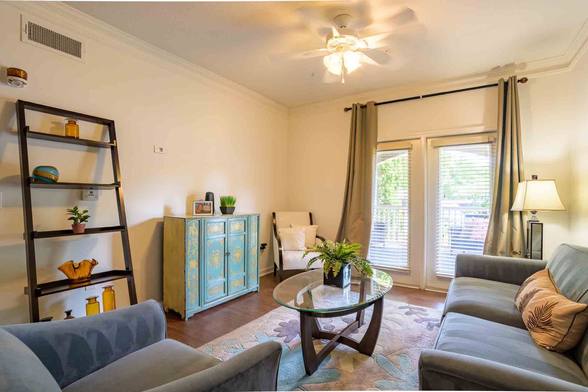 Living room: teal cabinet, gray sofa, glass table, wooden shelves, balcony access, tan curtains at Ansley Falls Apartment Homes in Charlotte, NC.