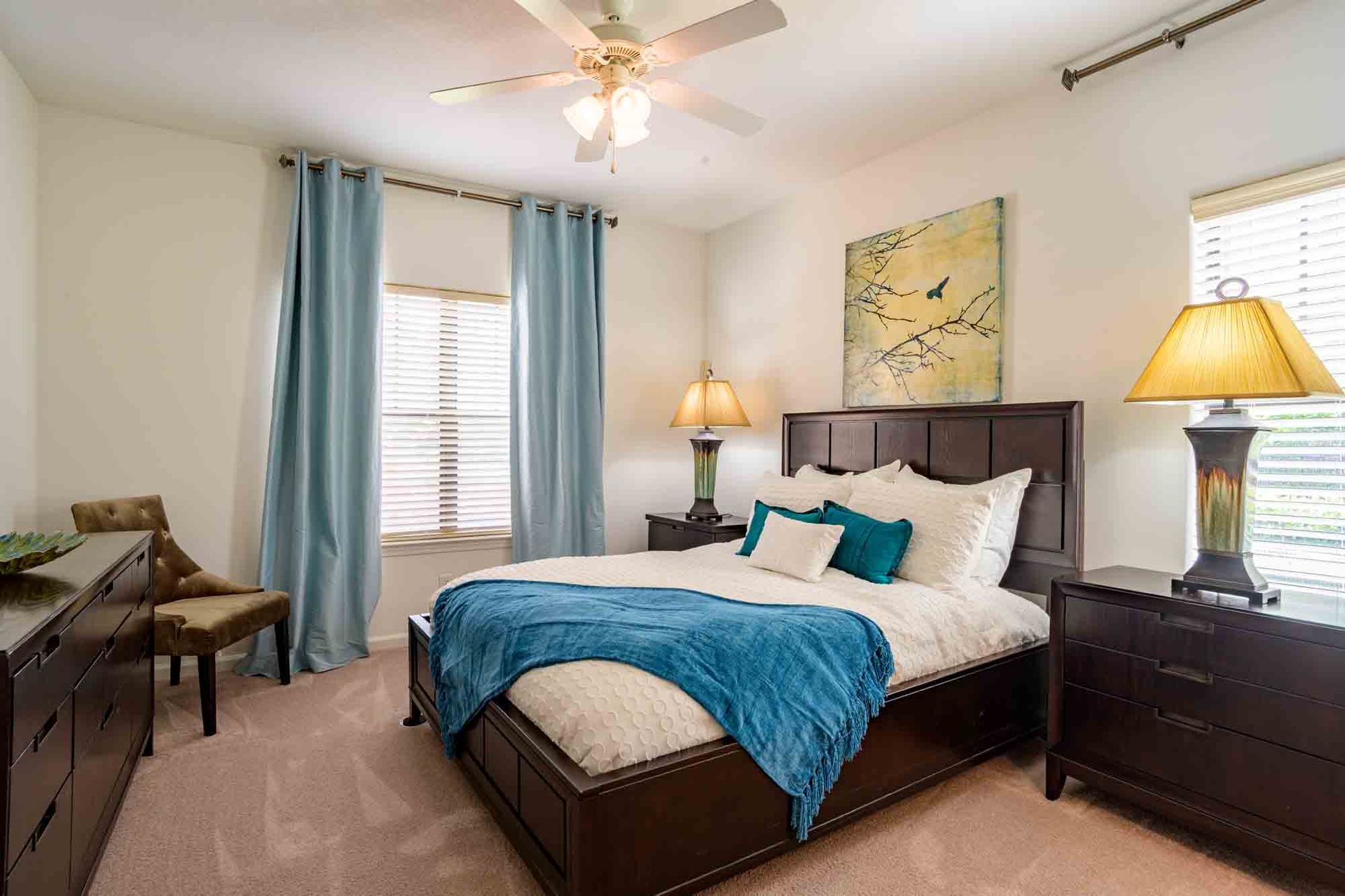 Bedroom with a dark bed frame, blue accents, lamps, and light-colored walls and carpet at Ansley Falls Apartment Homes in Charlotte, NC.
