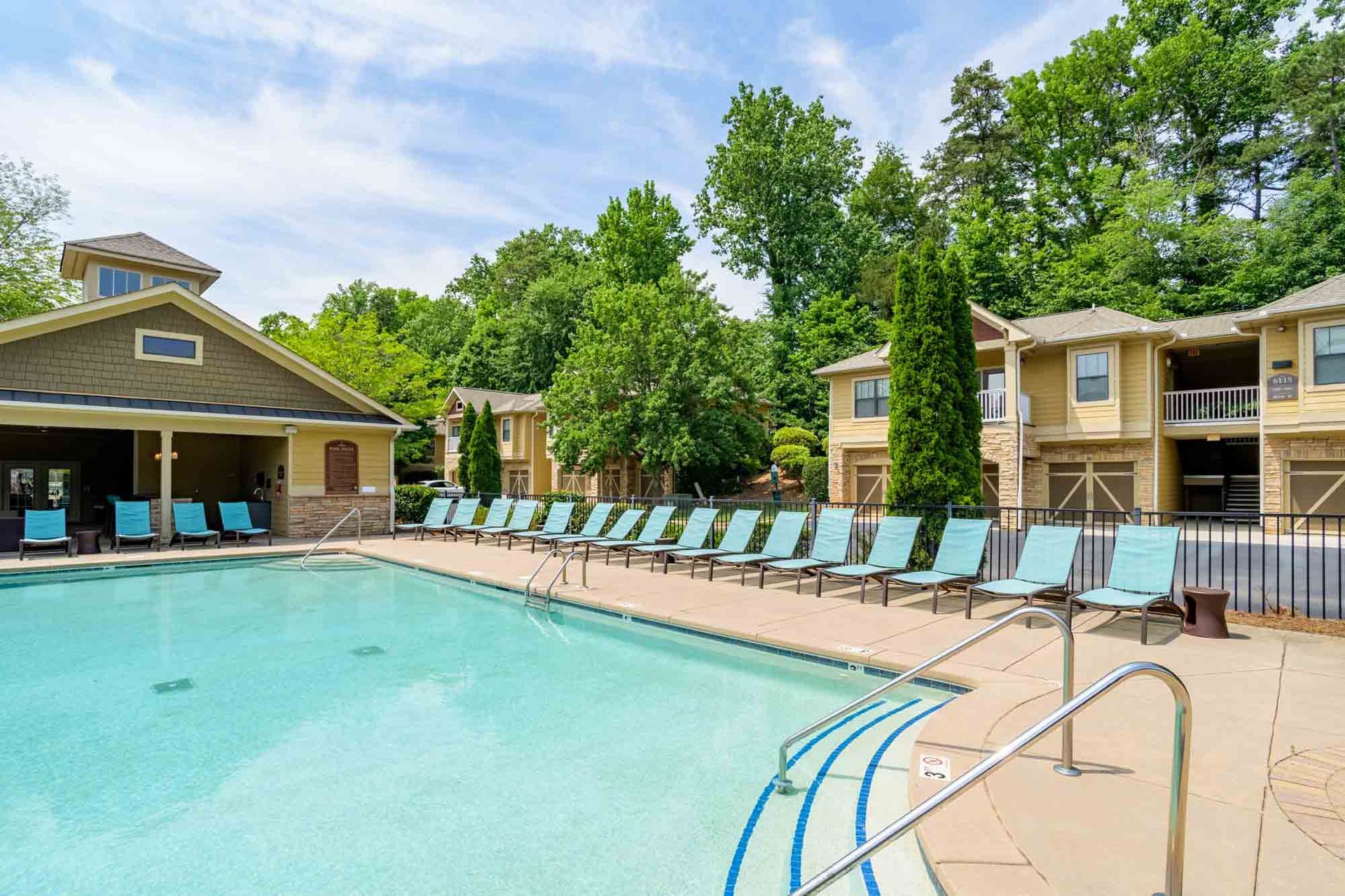 Pool with lounge chairs, buildings, and trees under a blue sky at Ansley Falls Apartment Homes in Charlotte, NC.