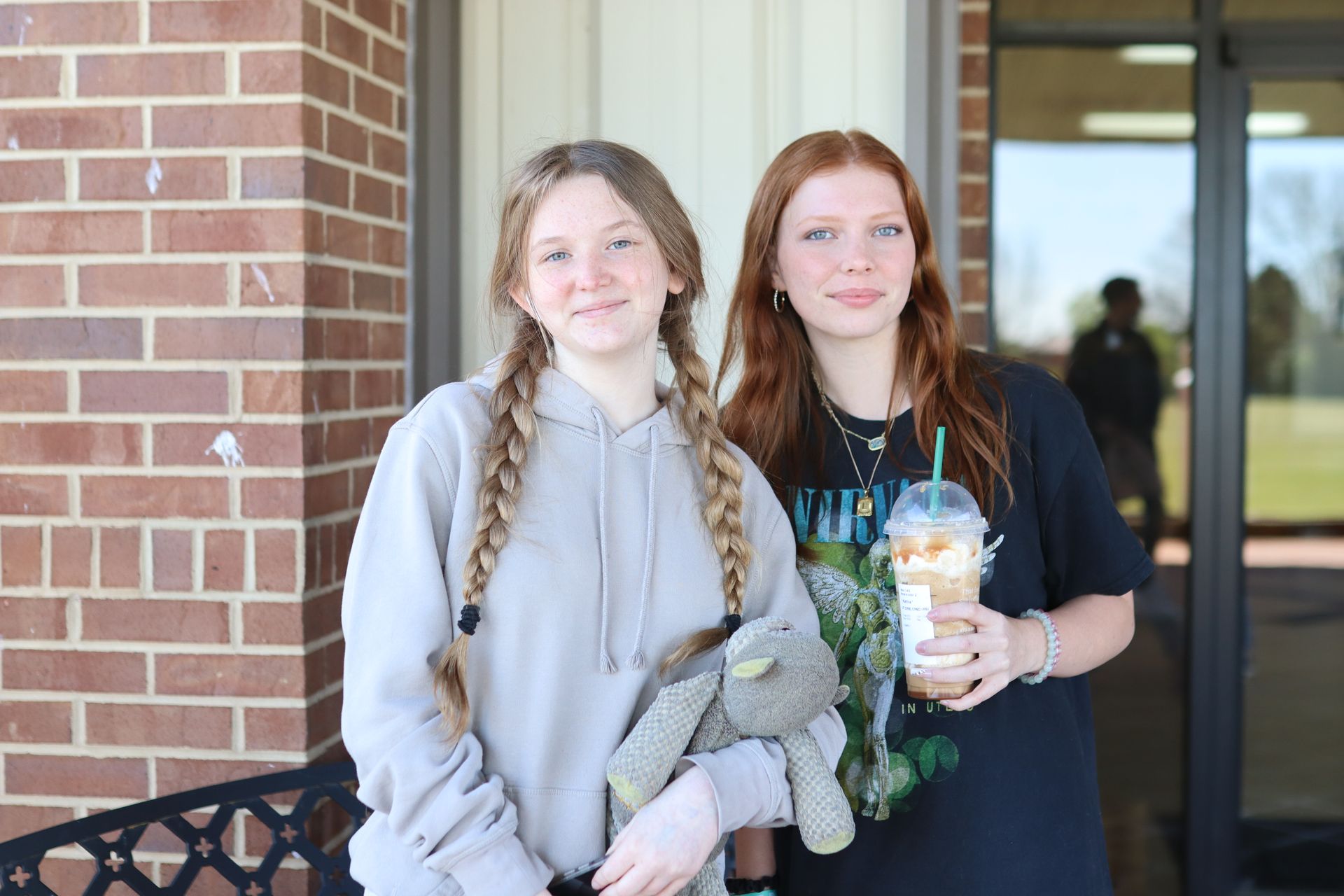 Two girls are standing next to each other in front of a brick building holding drinks and a stuffed animal.