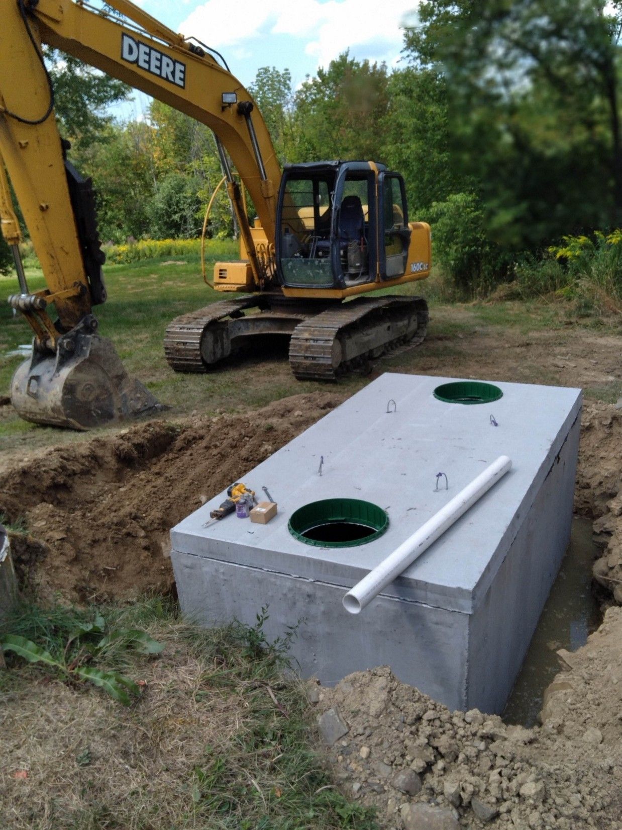 Excavator beside an underground utility tank with two green manholes at a dirt worksite