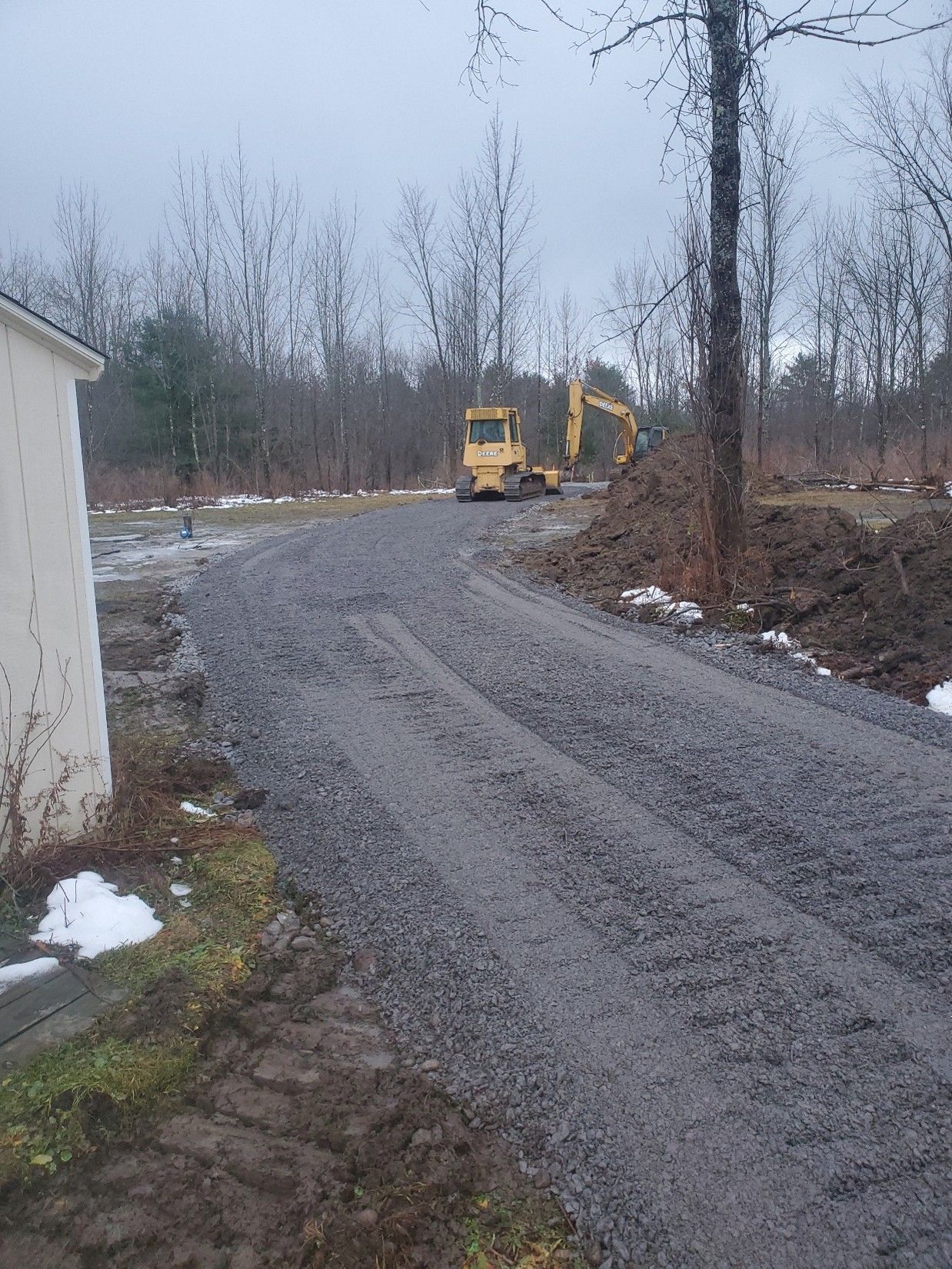 Gravel road curving past a white building, with a yellow excavator in a wooded area on a gray day