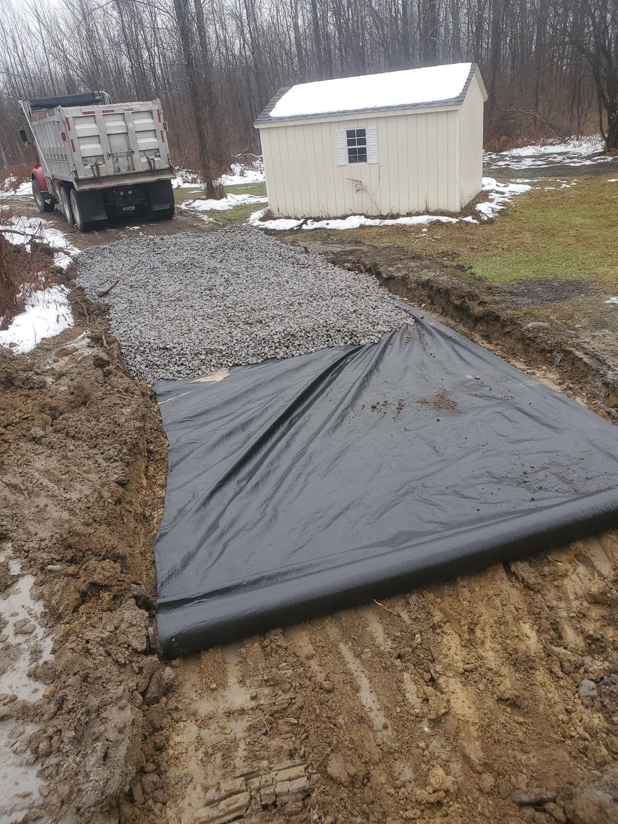 Muddy driveway with black tarp, gravel path, a dump truck, and a small shed in a snowy yard