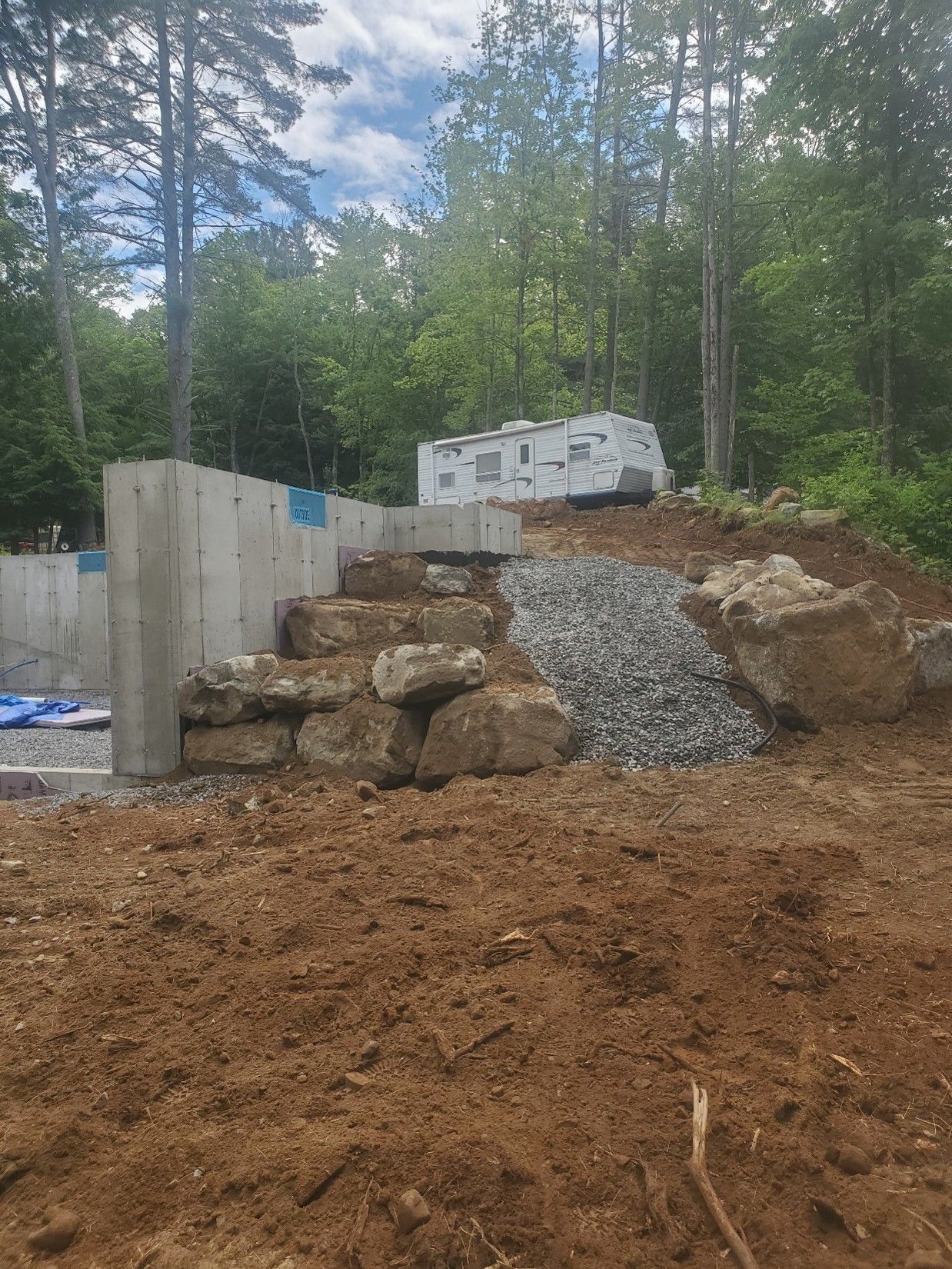 Gravel driveway under construction with retaining walls, large rocks, and a white trailer in a wooded area.