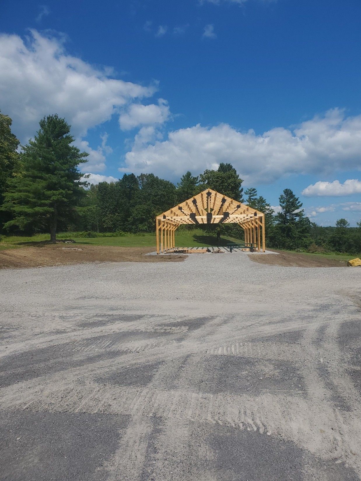 Wooden pavilion frame on a gravel lot with trees and blue sky in the background